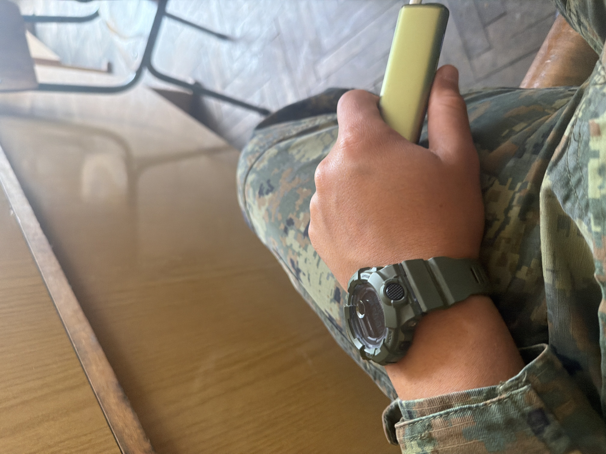 A man wearing a military-style camouflage uniform and a green G-Shock watch is sitting at a wooden table. He is holding a small, rectangular yellow device in his hand. The table and a chair are visible in the background. I have no information about the location of Dolna Mitropolia, Bulgaria.