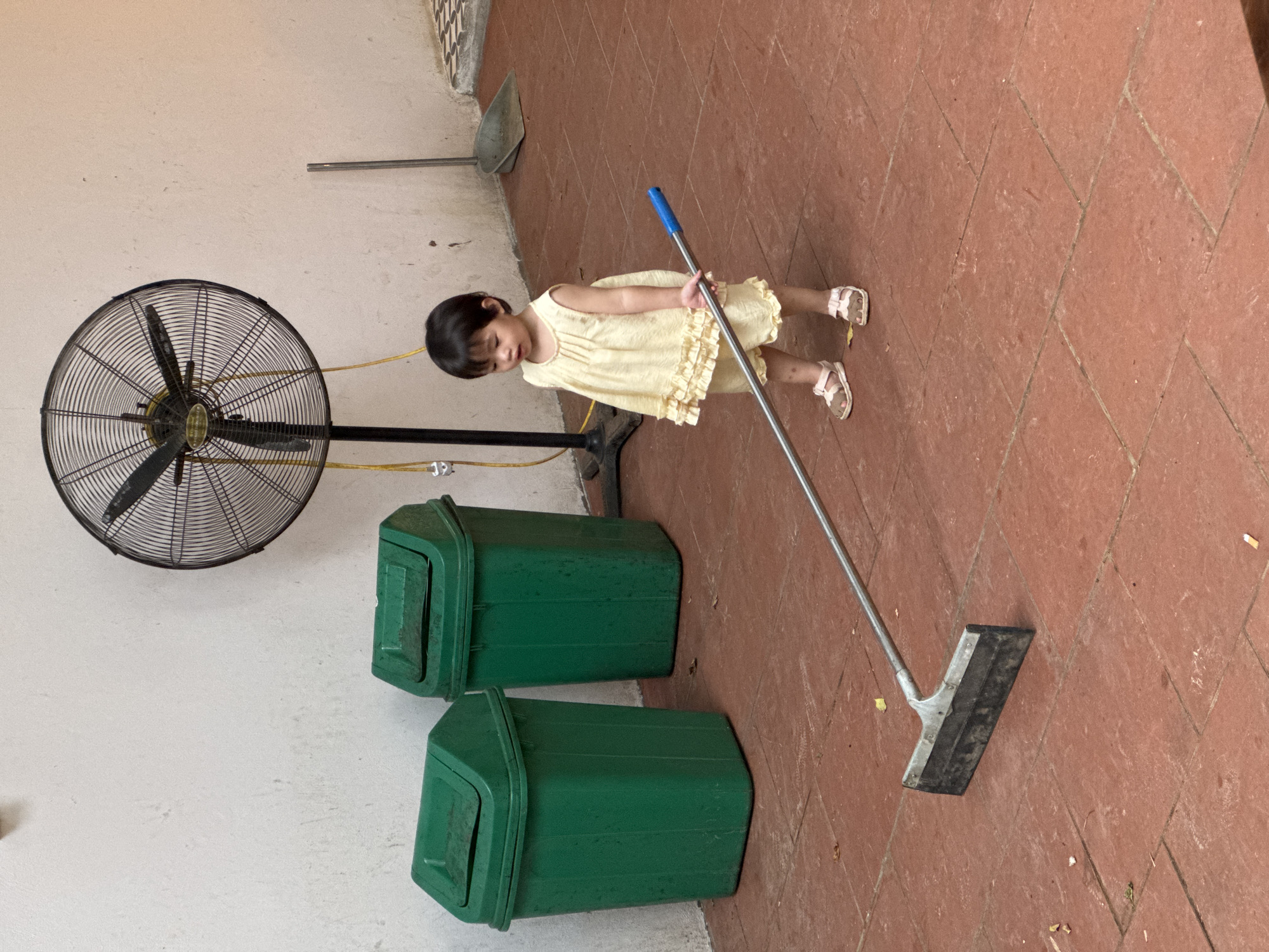In Cổ Lũng Commune, Vietnam, a young child, appearing to be around 2-3 years old, stands on a reddish-brown tiled floor, holding a floor squeegee with a blue handle. The child is wearing a light yellow ruffled dress and sandals. Behind the child, a large black oscillating fan with a yellow cord is mounted on a stand. To the left of the fan are two large green trash cans with lids. Further in the background, a dustpan and broom are propped against a wall. The overall environment suggests an outdoor or semi-outdoor space with a tiled floor and a simple, functional setting.