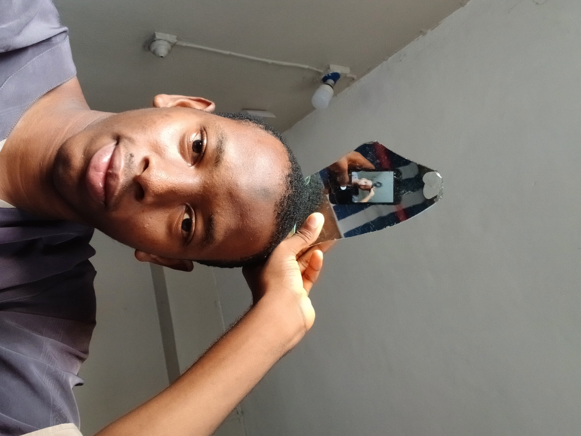 A close-up indoor shot shows a young Black man holding a shard of broken mirror to his right side. His face is turned towards the camera, and he appears to be taking a selfie with a mobile phone. The mirror reflects a part of the room. The location cannot be determined from the image. The ceiling has light fixtures. The background is a plain white wall. The man is wearing a gray shirt.
