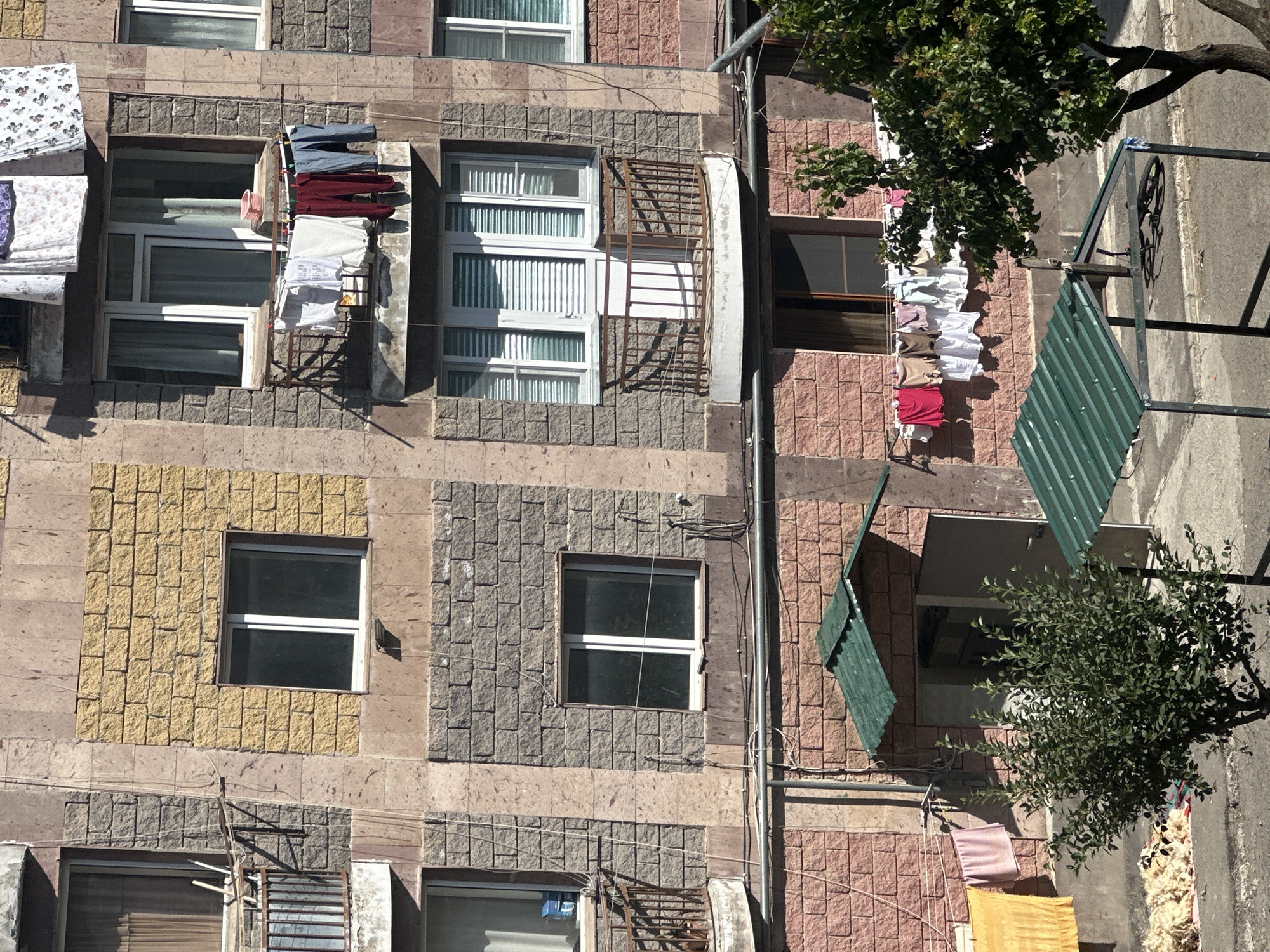 The image depicts the facade of an apartment building in Gyumri, Armenia, with laundry hanging on lines strung across balconies and windows. The building has a segmented design, with different sections clad in various materials, including textured yellow and grey stone-like panels, and lighter colored stone accents.
On the upper floors, several windows are visible, some with curtains. A balcony on the second floor from the top features laundry, including shirts and pants, drying on a clothesline. Above this, laundry is also seen hanging from an upper story.
The lower section of the building has red brick-like cladding. A small entrance with a green corrugated metal roof extends from this level. A bush is situated in front of this entrance, and some laundry, including a yellow towel and pink fabric, is hanging on a line to the left. Piles of light-colored material, possibly wool or bedding, are on the ground in front of the building.
A green corrugated metal canopy supported by metal poles is visible in the foreground, with a bicycle leaned against one of the poles. A tree with dense green foliage is to the right of the canopy, partially obscuring the building. The overall scene suggests a residential area where residents are drying their laundry outdoors.