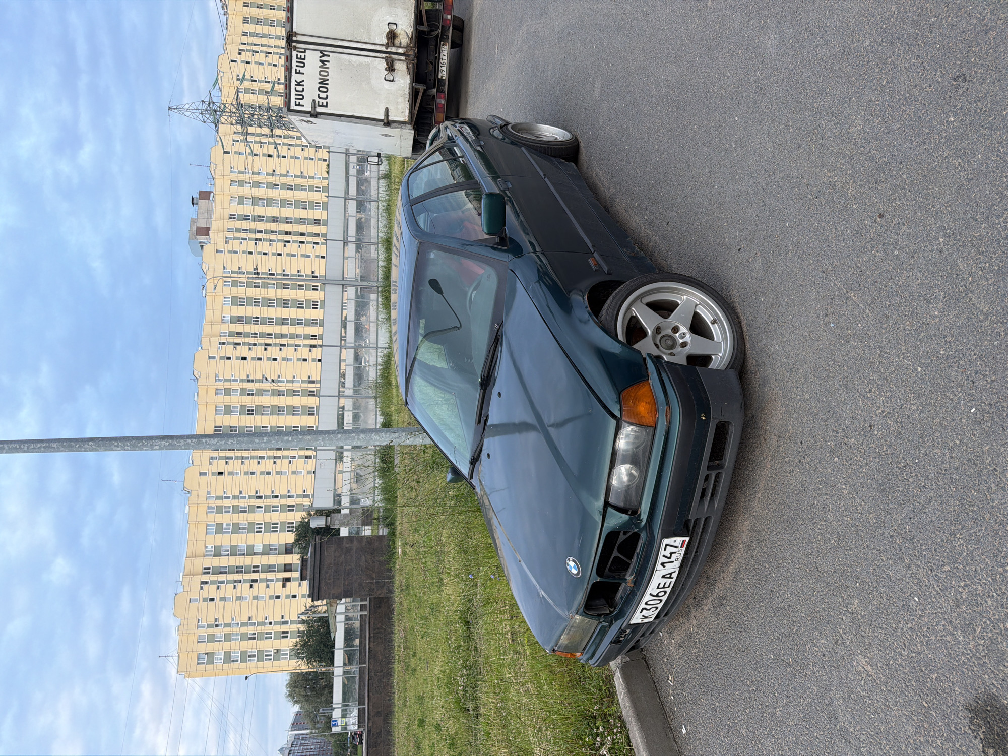A dark green four-door sedan with a custom license plate "K306EA 147 RUS" is parked on an asphalt road. The car has aftermarket silver five-spoke rims and a sun-damaged exterior. In the background, a large, pale yellow apartment building stands under a cloudy sky. A white truck with the text "FUCK FUEL ECONOMY" written on its rear door is also present. The scene appears to be in an urban environment, possibly an industrial or residential area.