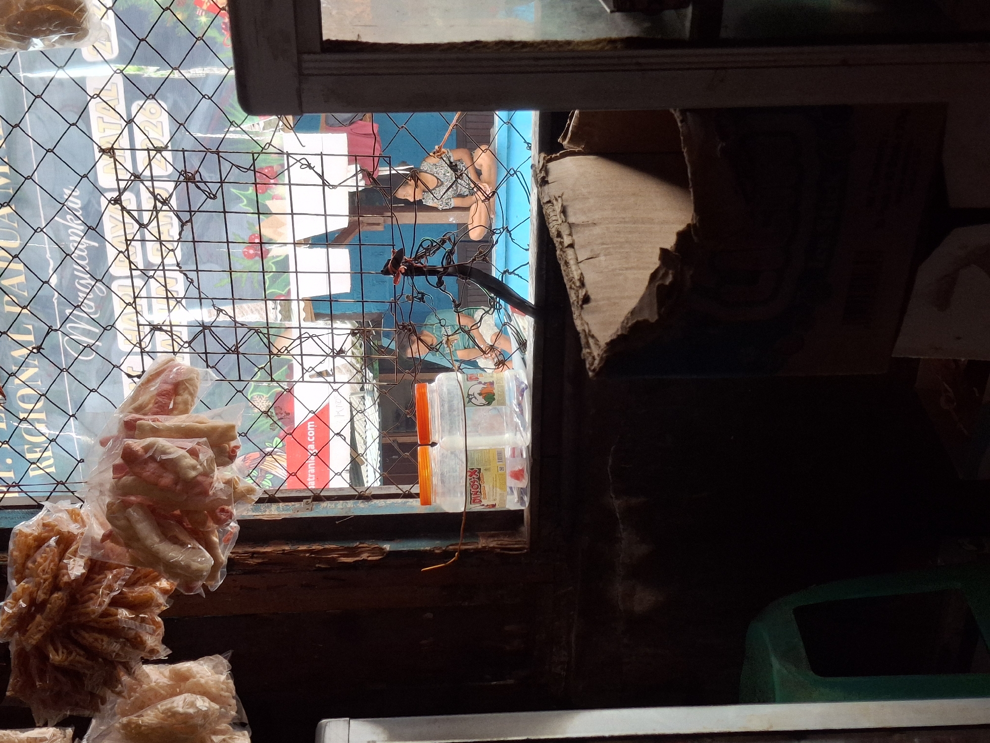 This is a medium shot image taken through a chain-link fence, focusing on a window of a shop in Biak, Indonesia. Inside, two children are visible. One child, on the left, is wearing a green t-shirt and sitting with their legs extended. The other child, on the right, is sitting cross-legged and wearing a patterned t-shirt. Both children are looking towards the right, away from the camera.
In the foreground, on a windowsill, are two clear plastic jars with orange lids. The jar on the left has a red and yellow label with text including "DINO-X" and an image of vegetables. The jar on the right also appears to contain a product. Behind the jars, and partially obscured by the fence, is a blue tarp or wall, and a banner with text. Part of the banner visible includes "AL PAPUA M...", "Moringankan", and "NATAL 2026". The url "satranilaga.com" is also visible on the banner.
Hanging from above, and also visible through the fence, are several plastic bags filled with food items that appear to be dried snacks or crackers. To the left of the window, part of a green plastic chair is visible. In the lower right corner of the image, a damaged cardboard box is in the foreground. The overall environment suggests an indoor market stall or shop.