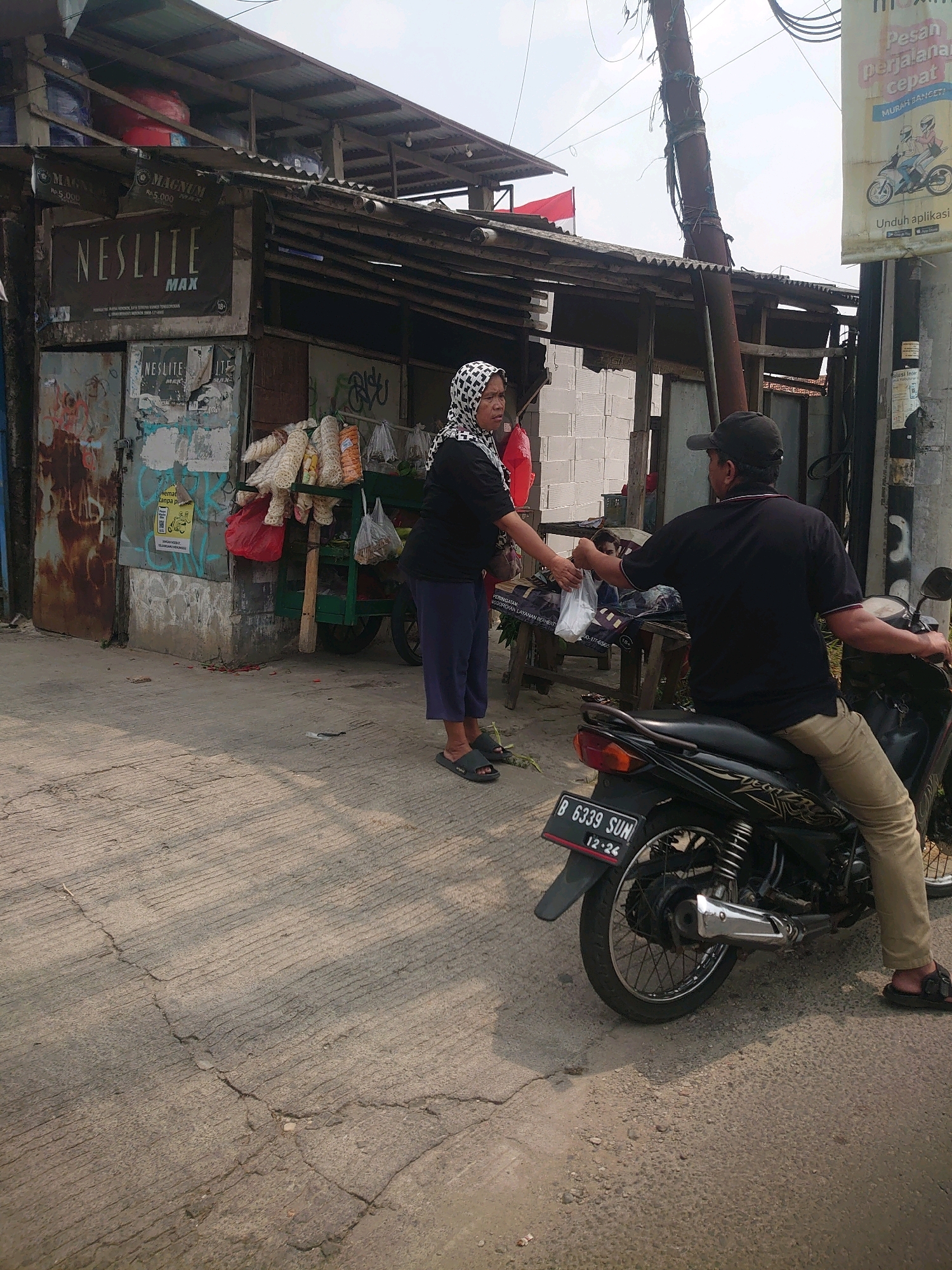 In Depok, Indonesia, an outdoor scene unfolds where a woman, wearing a black top, blue pants, and a black and white patterned headscarf, is handing a plastic bag to a man seated on a black motorcycle. The man, dressed in a black shirt and khaki pants, wears a dark baseball cap. The motorcycle has a license plate that reads "B 6339 SUN 12-26". They are standing near a roadside stall constructed with corrugated metal roofing supported by wooden poles, with a partial white brick wall visible behind it. The stall displays various items, including packaged snacks or food items hanging from a rack, and a red bag. To the right of the motorcycle, a utility pole with wires is prominent. The ground is paved with concrete, showing cracks and a textured surface. In the background, a tall, yellow advertisement poster is attached to a pole. The sky above is partly cloudy.
