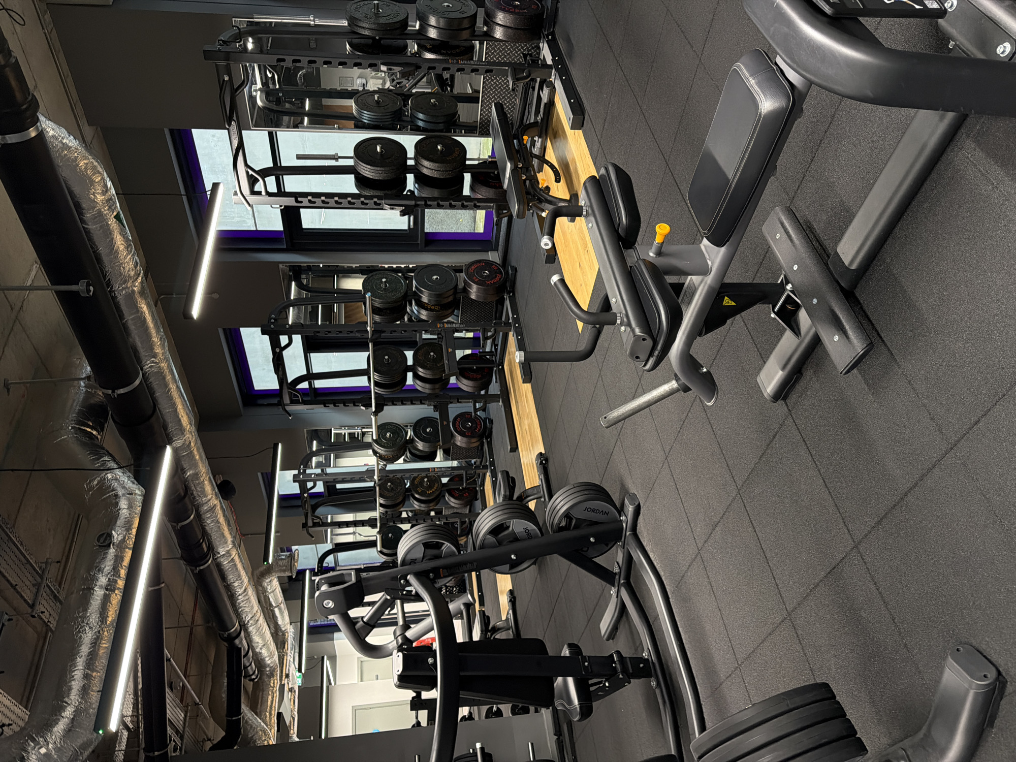 The image depicts the interior of a gym. The gym is equipped with weightlifting equipment, including weight racks filled with various weight plates. There is also exercise machinery. The floor is covered in dark gray rubber tiles. The ceiling features visible ductwork. The location is likely in London, United Kingdom. No people are visible in the image.