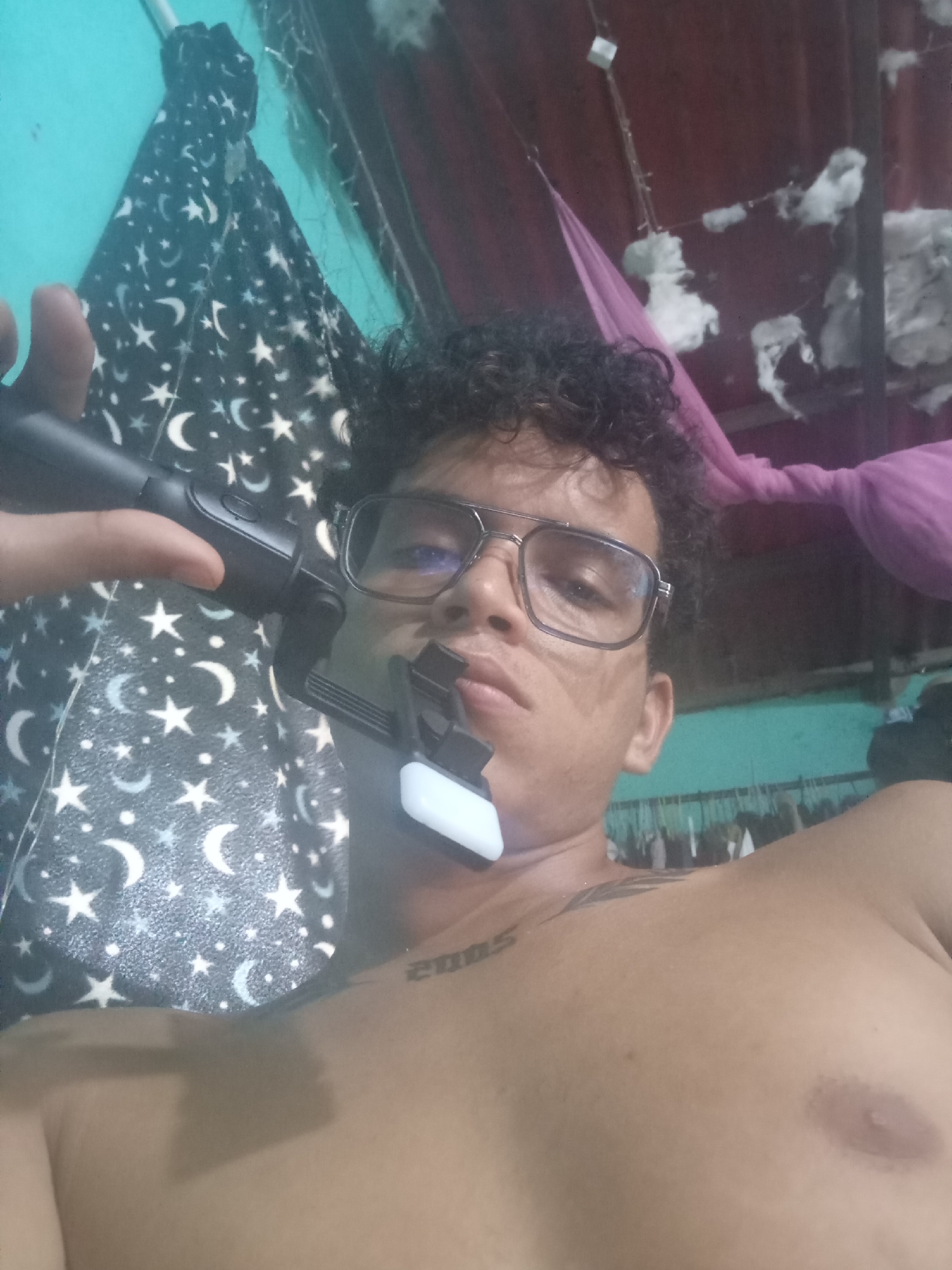 The image shows a person holding a selfie stick, with the camera pointing towards their face. The person is wearing glasses and has curly hair. There is a black and white patterned fabric with stars and moons in the background, and some pink fabric is visible further back. The person has a tattoo on their upper chest. The environment appears to be indoors, possibly a bedroom, with some items hanging on a wall in the background. The lighting is dim. The image does not provide any specific clues to determine the location within the city of Altagracia, Venezuela.