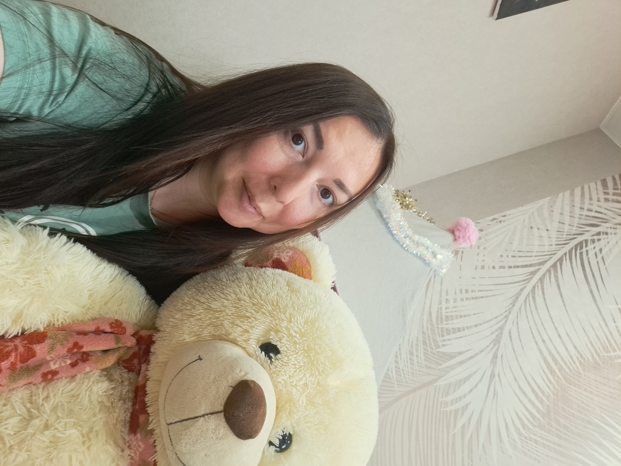A woman with long, dark brown hair is lying down, her head tilted towards the camera. She is wearing a light green t-shirt. A large, fluffy teddy bear is positioned in front of her. The bear has a light-colored fur, a brown nose, and a floral-patterned scarf around its neck. The background includes a wall with a white and light grey tropical leaf pattern. There is also some Christmas decoration hanging near the wall. The location is Chaikovsky, Russia.