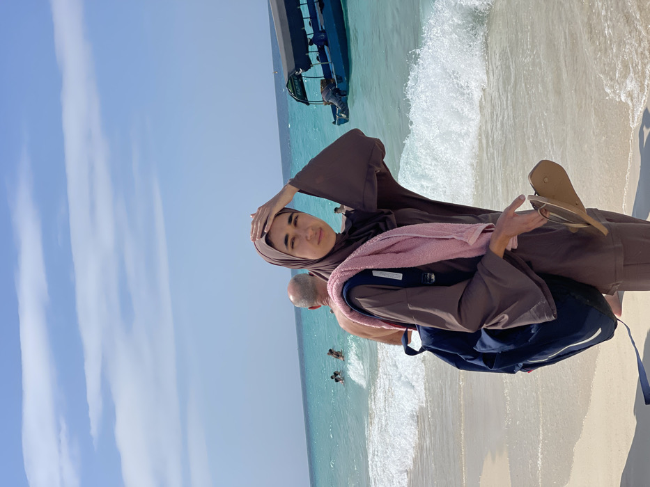 A young adult female stands on a sandy beach in Zanzibar, Tanzania. She wears a brown hijab and a matching long-sleeved tunic and trousers. A dark blue backpack rests on her left shoulder, draped with a light pink towel. She holds light brown flip-flops in her right hand, while her left hand is positioned on her head. She faces the viewer with a neutral expression. Behind her, partially submerged in the turquoise water, a bald adult male with his back to the camera observes the sea. Several other individuals are visible swimming or wading further offshore. To the right, a blue boat with a canopy, labeled "KAZIMOTO" on its side, and an outboard motor is anchored or drifting in the water, carrying multiple passengers. White foam from breaking waves surrounds the main subject's feet. The sky above is clear blue with sparse white clouds.