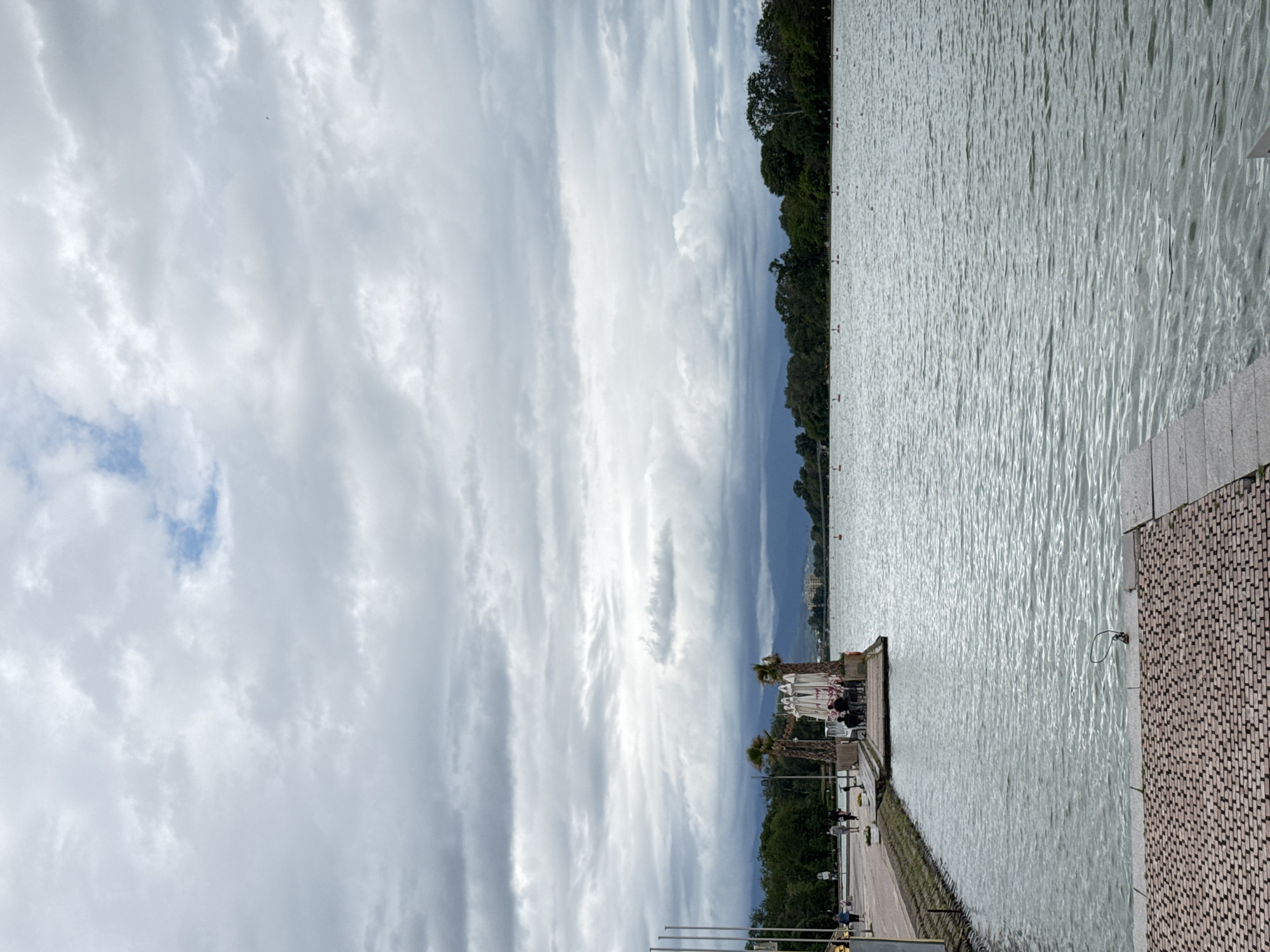The image depicts a scene along the Rowing Canal in Plovdiv, Bulgaria. The sky is predominantly covered in gray and white clouds, with a small patch of blue visible. A line of trees and vegetation borders one side of the water. A paved walkway runs alongside the canal, with people strolling and sitting at outdoor cafes under white umbrellas. Palm trees are interspersed along the walkway. The canal water is rippled, reflecting the cloudy sky above. A textured, paved area is in the foreground. The image is oriented sideways.