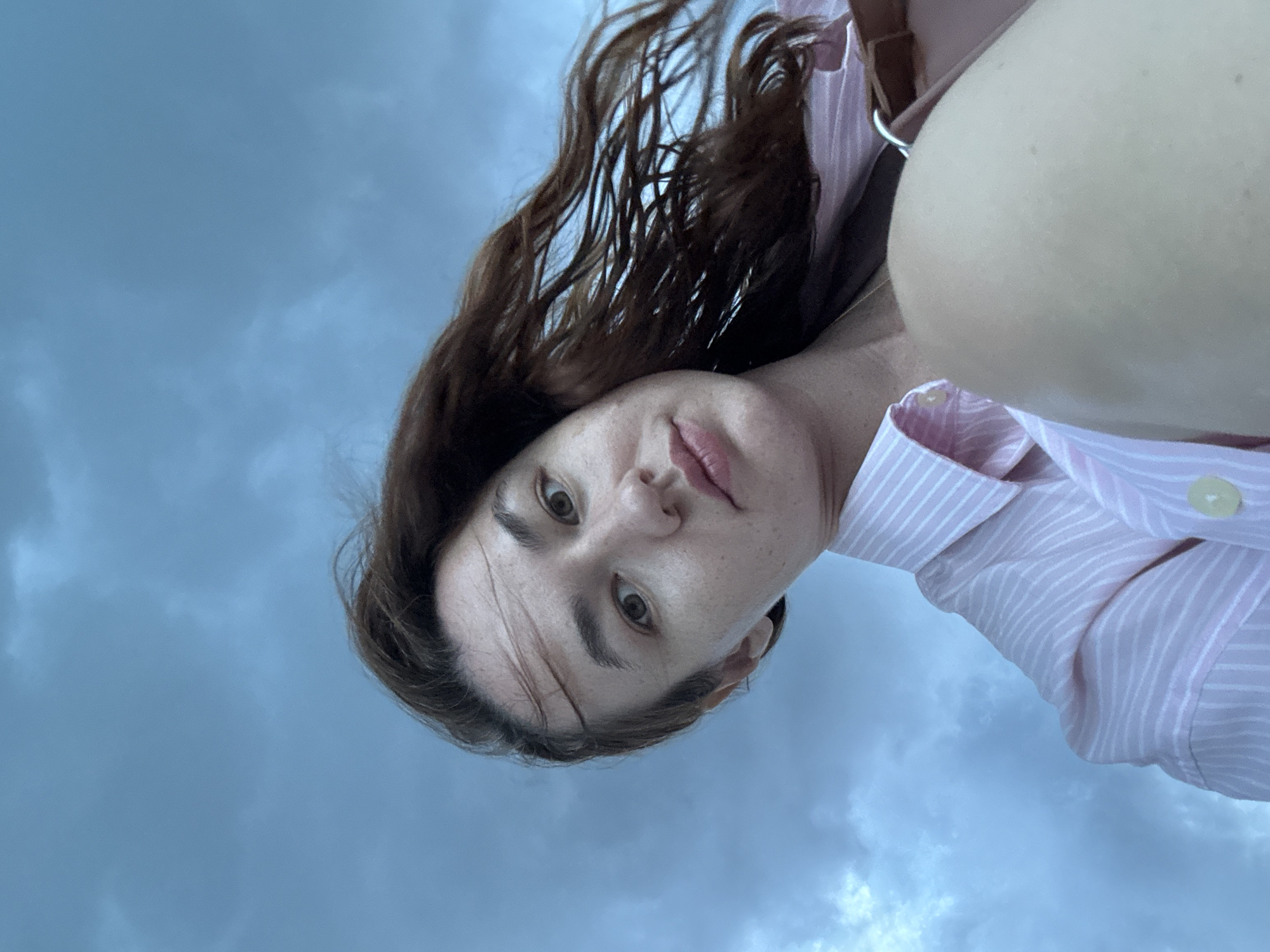 A low-angle, medium shot captures a young woman's face as she looks directly at the camera, her head tilted slightly upwards against a backdrop of a cloudy, blue sky. Her dark, wavy hair is blown to the right by the wind, obscuring part of her face and body. She is wearing a pink and white striped button-up shirt with the collar visible. The lower right portion of the image shows her bare knee and a glimpse of a brown strap, possibly from a bag. The overall lighting suggests an overcast day, and the scene appears to be outdoors. The location is Baan Chaweng Noi, Thailand.