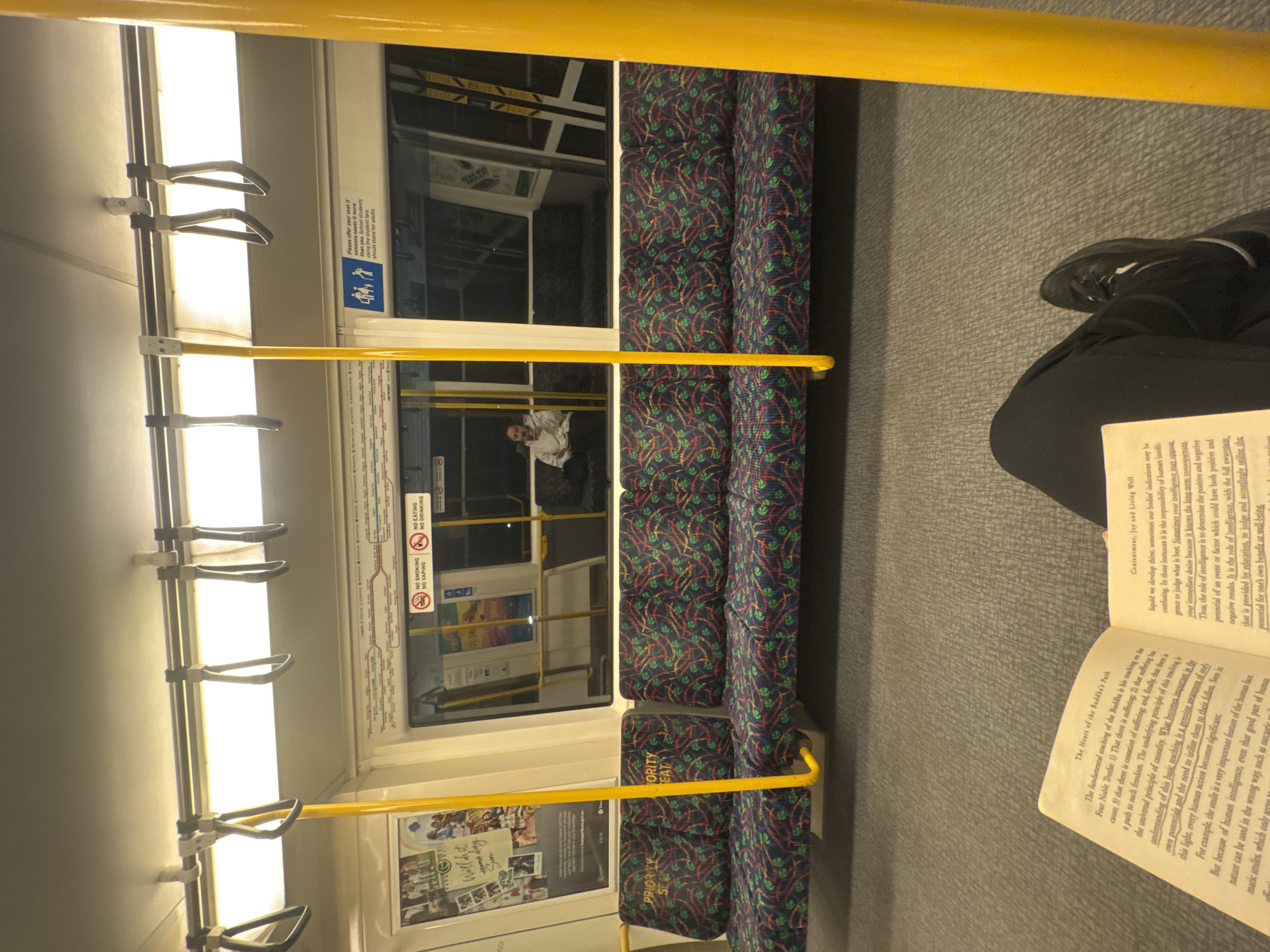 An interior view of a Perth public transport train carriage, likely a Transperth train, shows the reflection of the surrounding environment in the windows. The seats are upholstered in a dark fabric with a repeating pattern of red, green, and blue motifs. Yellow poles, commonly found in public transport for passengers to hold onto, are visible. In the foreground, an open book rests on the lap of a person wearing black pants and a black sneaker. The book's pages contain text, with some passages highlighted. Through the windows, the reflection shows other passengers, one individual in particular is seated and looking at their phone. Another section of the window displays signage indicating "NO SMOKING," "NO VAPING," and "NO DRINKING." A sign on the seats reads "PRIORITY SEAT." The overall environment suggests a typical commute on public transportation.