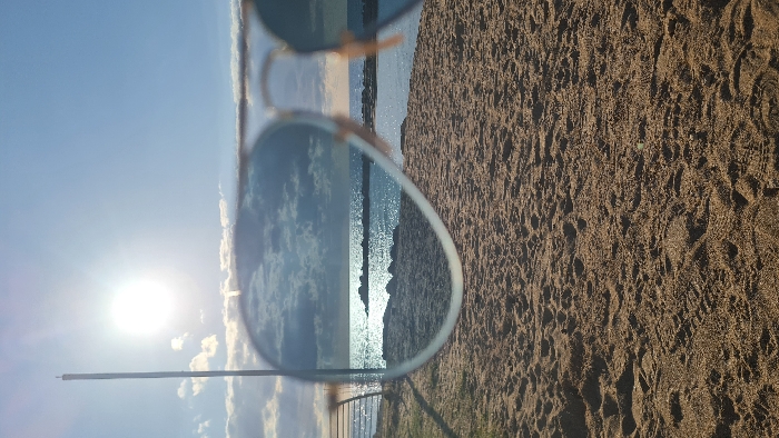 The image presents a daytime scene on a sandy beach in Nahariyya, Israel. The primary subject is a pair of gold-rimmed, aviator-style sunglasses positioned in the immediate foreground, partially obscuring the view. The reflective lenses of the sunglasses capture an image of a cloudy sky above the Mediterranean Sea, with glinting sunlight on the water's surface and multiple rock breakwaters visible in the distance. Above the sunglasses, the actual sky is bright blue with scattered white clouds, dominated by a radiant sun in the upper left quadrant. A tall, slender, light-colored pole extends vertically from the left side of the frame, partially visible against the sky. The lower half of the image is comprised of light brown sand, showing numerous footprints. The edges of the scene visible directly, not through the sunglasses' reflection, reveal the sandy beach extending to the water, where sun glint and rock breakwaters are also discernible. No individuals are present within the depicted landscape.