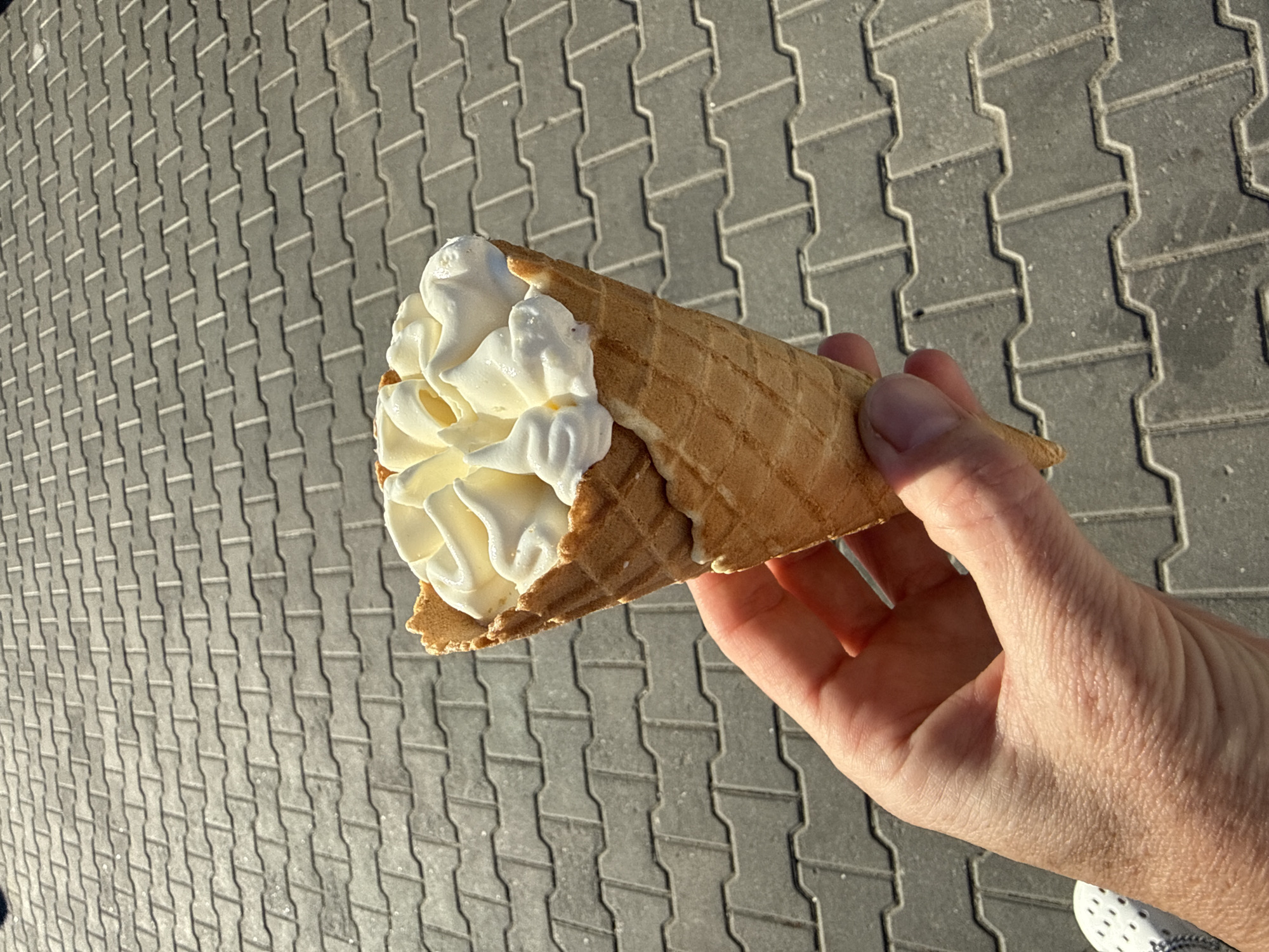 A hand holds a vanilla ice cream cone against a backdrop of grey interlocking pavers. The ice cream is swirled and appears to be melting slightly. The background suggests an outdoor environment with artificial lighting.
