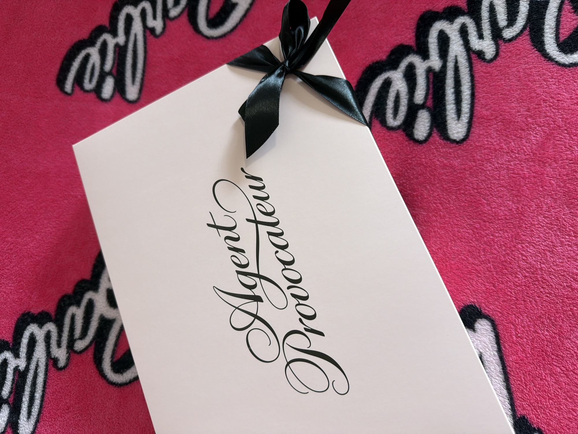 A close-up studio shot features a white Agent Provocateur box, tied with a black satin ribbon, set against a pink fuzzy Barbie-themed blanket. The box is positioned at an angle, displaying the brand name in cursive black lettering. The ribbon is tied in a bow at the upper edge of the box. The background is pink, covered in black and white "Barbie" text. The overall setting is indoors, with controlled lighting. The location of the scene is unknown.