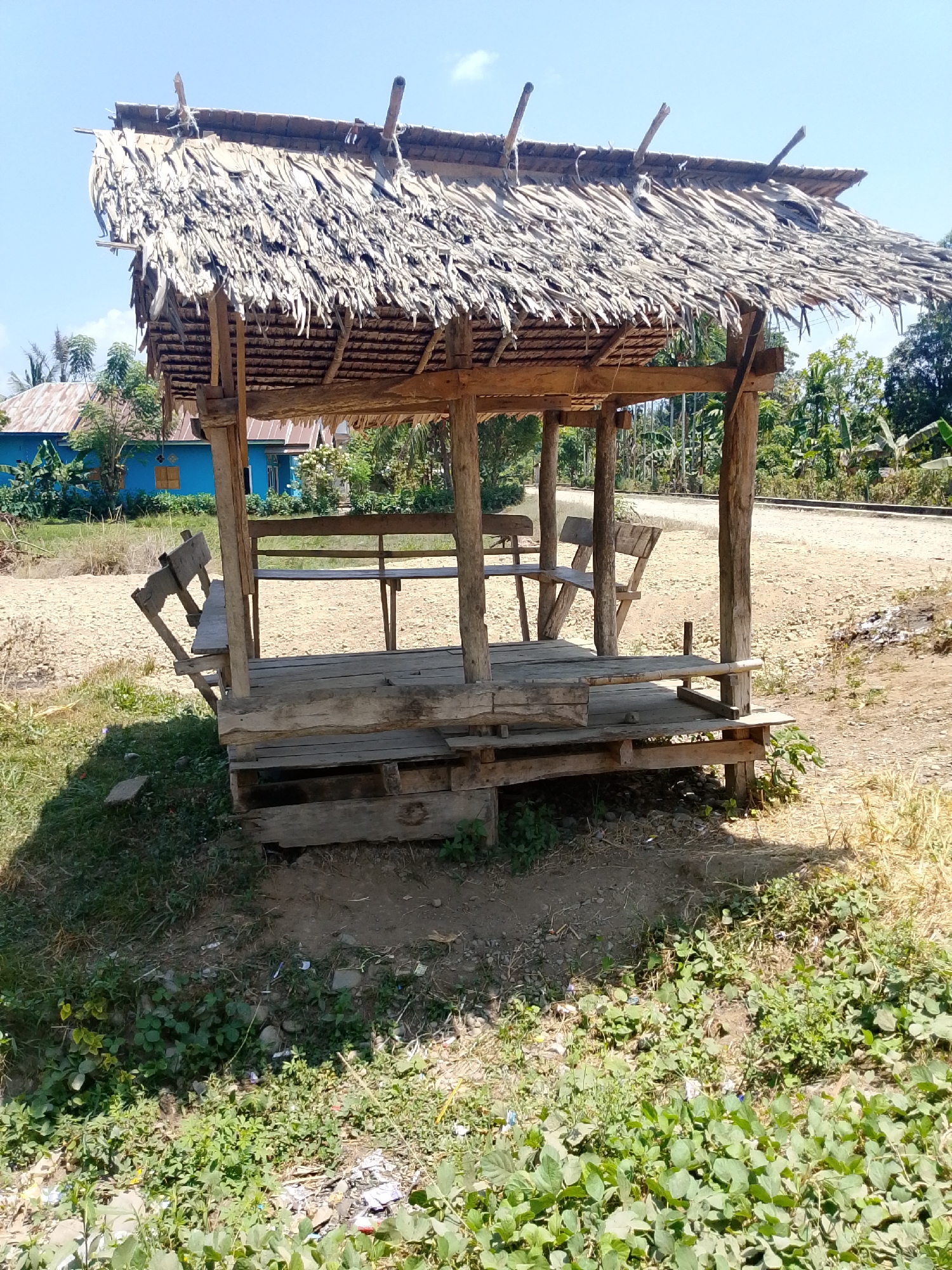 A rustic wooden structure with a thatched roof stands under a clear blue sky in Se'pon, Indonesia. The structure, likely a shelter or pavilion, is built with rough-hewn wooden posts and beams. The roof is thatched with dried palm leaves, giving it a textured, natural appearance. Inside the structure, wooden benches are visible, suggesting it is a place for seating or resting. The ground around the structure is a mix of dry earth, gravel, and sparse vegetation. In the background, a bright blue building with a dark roof is visible, along with trees and foliage, indicating a semi-rural or suburban setting. A dirt road runs behind the structure, bordered by more trees. The overall scene depicts a simple, open-air structure in a natural, outdoor environment.