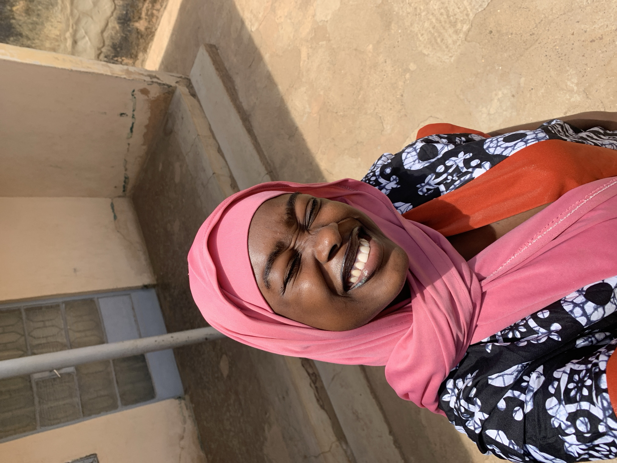The image is an eye-level shot of a woman with dark skin wearing a pink hijab. Her eyes are closed and she is smiling broadly, revealing her teeth. She is wearing a black and white floral print top with an orange shawl. The background shows a building with a window, and a concrete surface. The image was captured in Rafin Par, Nigeria, based on the prompt.