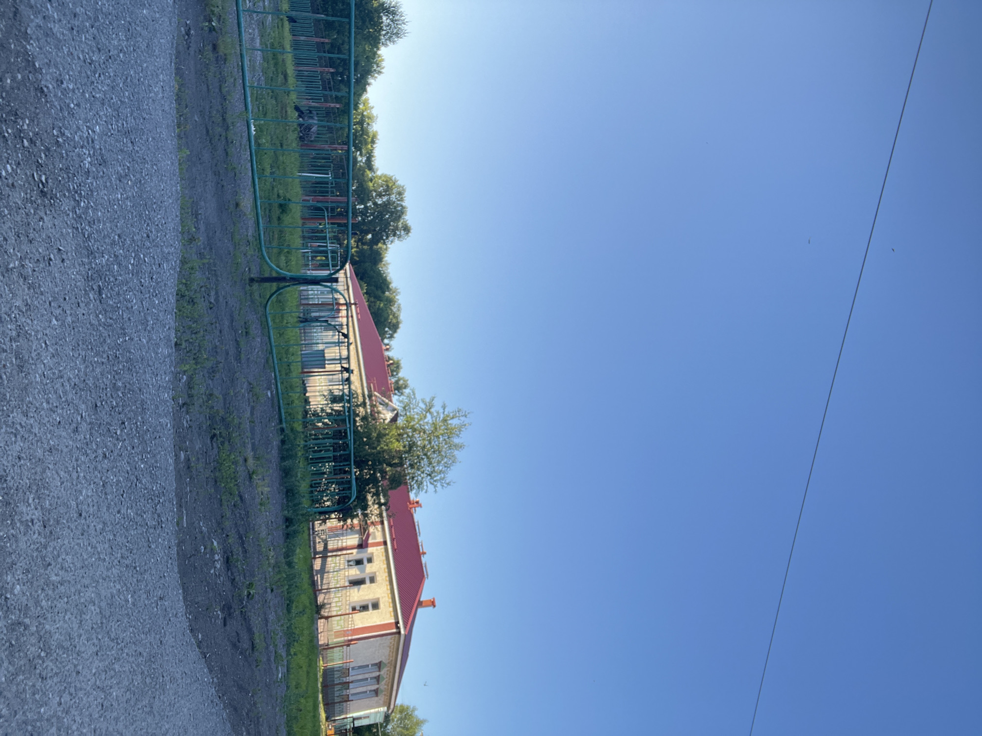 This image depicts a residential building with a red roof, possibly a school or administrative building, in Kiselyovsk, Russia. The building is light-colored with red trim and has multiple windows. It is partially obscured by a green fence. To the left of the building, there is a gravel pathway and some green vegetation. The sky is a clear, bright blue, suggesting a sunny day with good weather. The sun appears to be high in the sky, indicating it is likely midday. There is a thin black wire stretching across the sky. No people are visible in the image. The overall impression is one of an ordinary day in a semi-urban or rural setting.