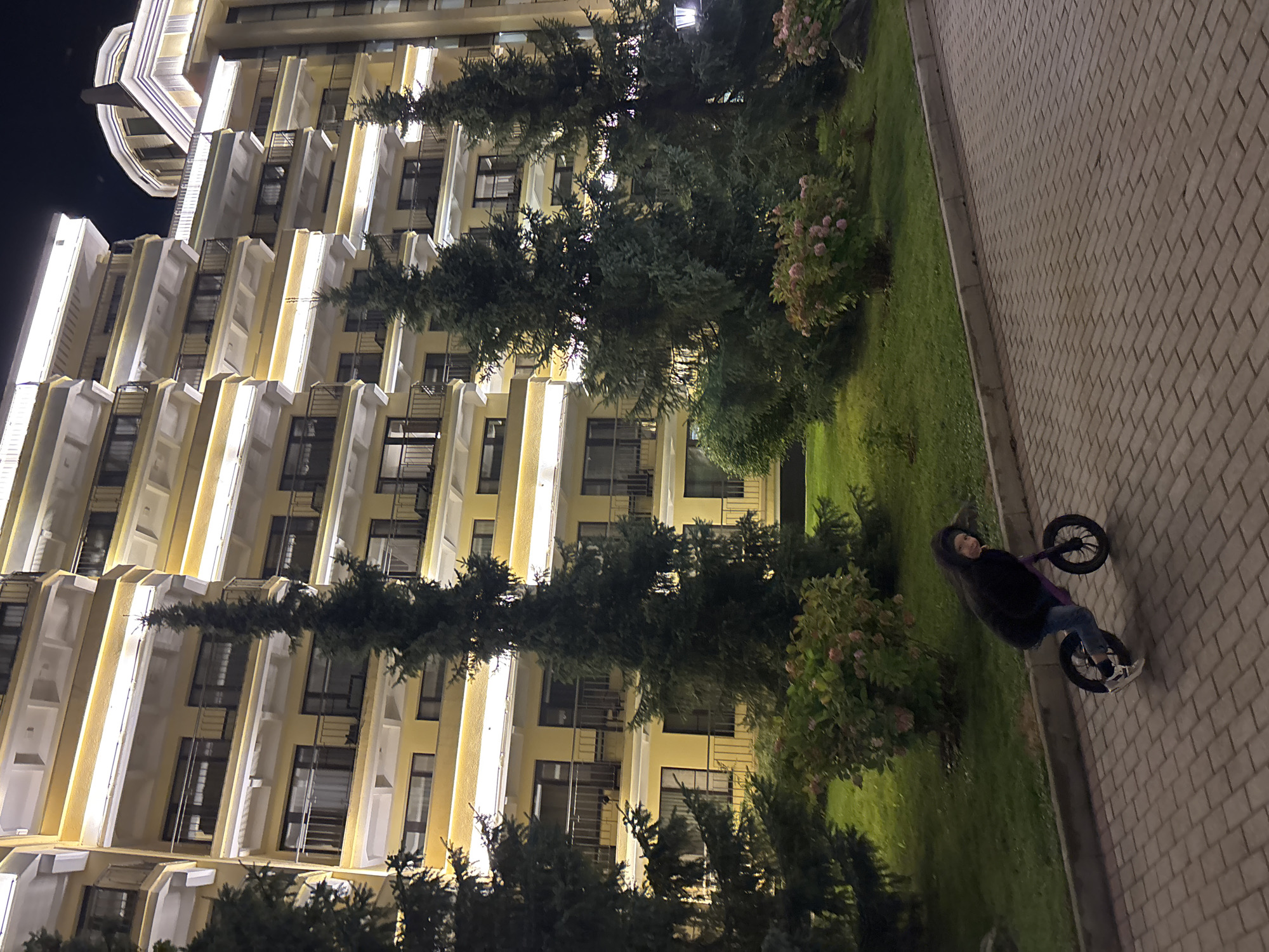 A child with dark hair and a black hooded jacket rides a small purple balance bike on a paved walkway in front of a multi-story yellow building at night. The building is illuminated by warm yellow lights along its edges and balconies. Tall evergreen trees and bushes with pink flowers are visible on a grassy area between the building and the walkway. The walkway is made of light brown rectangular pavers. The child is leaning forward, appearing to be in motion. The overall scene suggests an urban or suburban residential area. The location is identified as Рождествено, Russia.