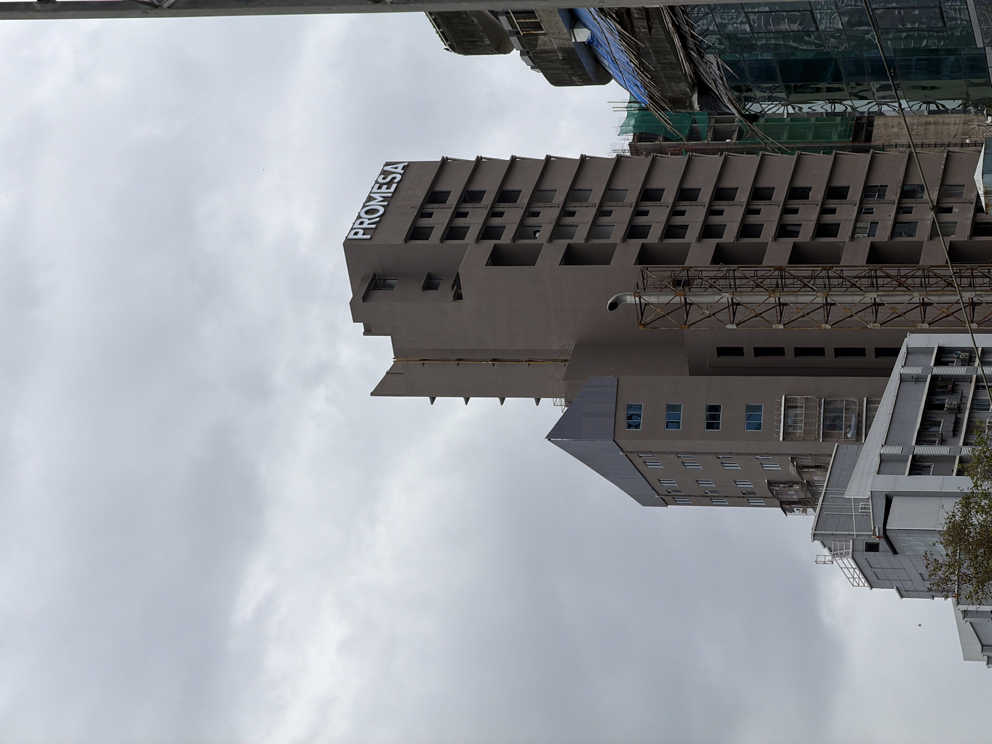 The image shows a cluster of buildings in Mumbai, India, under a cloudy sky. The prominent structure is a modern, multi-story building with a dark grey facade, topped with a large, white illuminated sign that reads "PROME SA." Several floors of this building have empty window frames, suggesting it is either under construction or recently completed. Adjacent to this building on the left is a lower, lighter grey building with a distinct angular roofline and visible windows. To the right, another building, partially obscured, appears to be under construction, with scaffolding and green netting visible. The overall scene depicts an urban environment with active construction and architectural development.