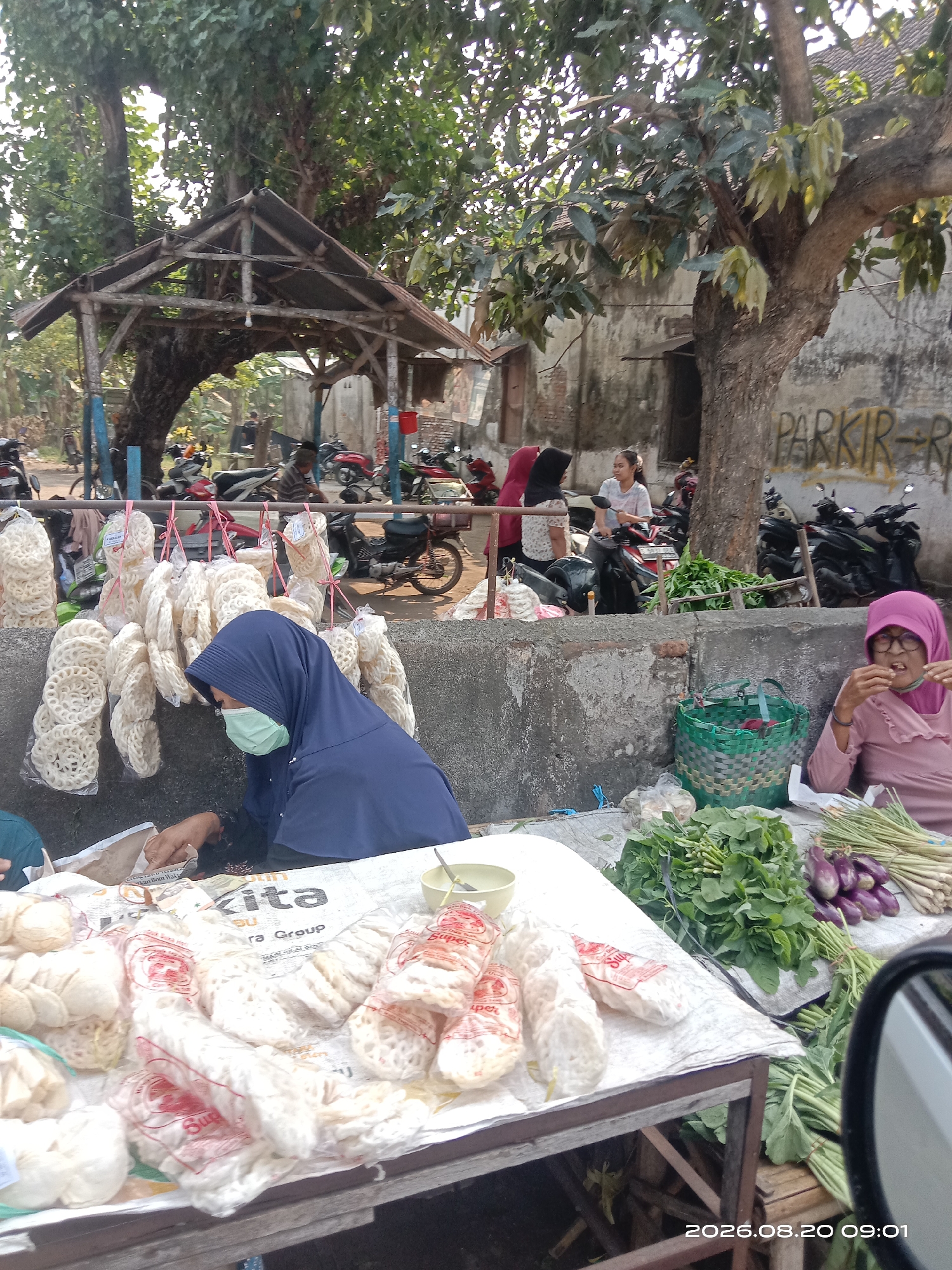 The image depicts an outdoor market scene in Gudo, Indonesia, likely a roadside market given the presence of parked motorcycles. In the foreground, a wooden table displays various food items, including what appear to be packaged crackers, fried snacks, and fresh produce such as leafy greens, lemongrass, and eggplants.
A woman wearing a blue hijab and a light green face mask is seated behind the table, interacting with something on the table. To her right, another woman, wearing a pink hijab and glasses, is seated and eating something.
In the background, several motorcycles are parked, suggesting this is a transportation hub or a place where people gather. Behind the market stalls, there is a rustic wooden shelter structure with a corrugated metal roof, and further back, a building with a faded "PARKIR" (parking) sign is visible. Trees with dense foliage provide shade over the market area. The overall impression is of a bustling, informal marketplace where daily commerce takes place. The timestamp "2026.08.20 09:01" is visible in the lower right corner of the image.