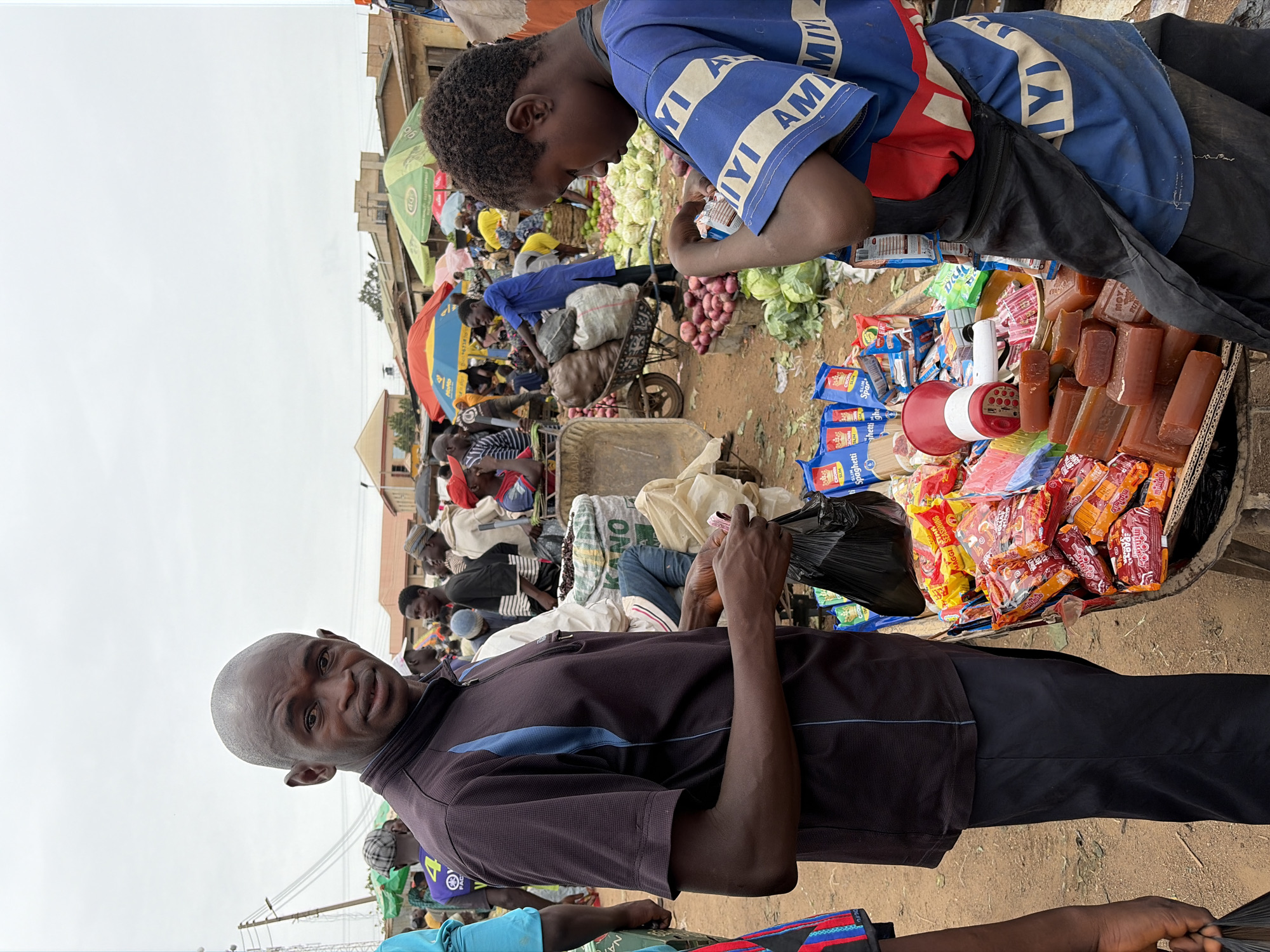 The image captures a bustling market scene in Gura Top, Nigeria. A man in the foreground, wearing a dark short-sleeved shirt and dark pants, holds a black plastic bag and looks directly at the camera with a smile. Behind him, a young person in a blue shirt with white lettering leans over a display of goods for sale. The display is on a table or cart and includes food items like packets of spaghetti, bouillon cubes, candy, and what appear to be bars of brown soap or similar products. A red and white megaphone is also visible on the display. In the background, a crowded marketplace unfolds with vendors and shoppers. Several individuals are gathered around a wheelbarrow filled with what looks like onions. Colorful umbrellas provide shade. The environment appears dusty or earthy.
