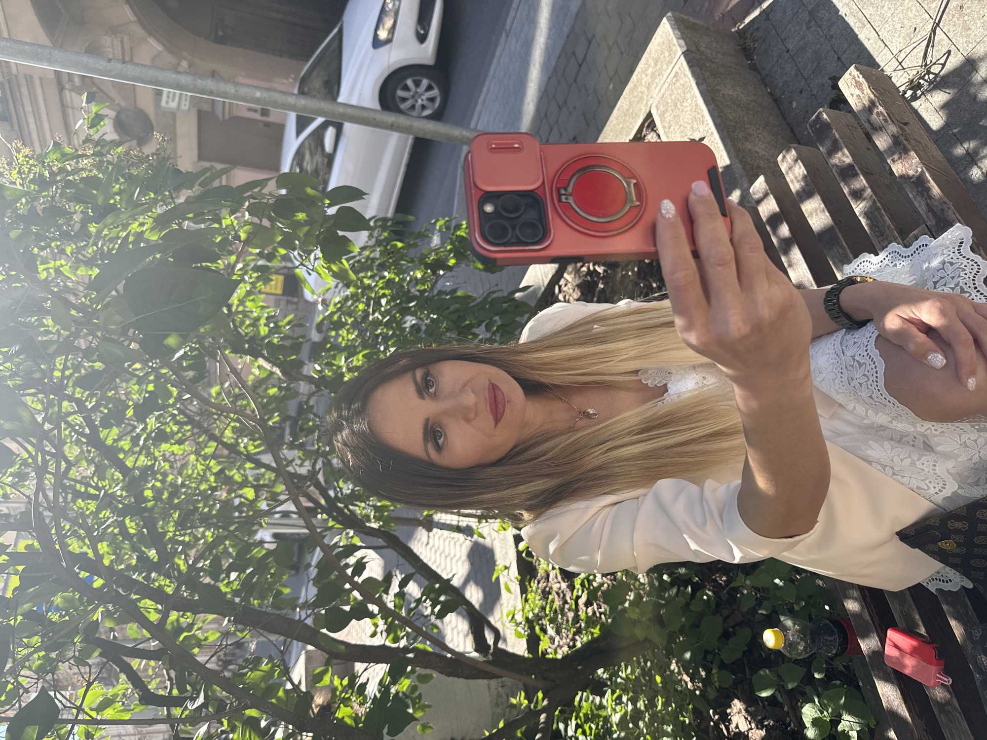 A woman with long blonde hair is sitting on a wooden bench in what appears to be a city street in Saint Petersburg, Russia, based on the architectural style of the building in the background and the car model. She is holding up a red smartphone to take a selfie. She is wearing a light-colored blazer over a white lace top and a delicate necklace. A bottle of dark liquid and a red object are on the bench next to her. Sunlight filters through the leaves of a tree in the foreground, casting dappled shadows on the scene. A white car is visible in the background on the street, and a lamppost stands to the right. The overall impression is a casual outdoor setting on a sunny day.