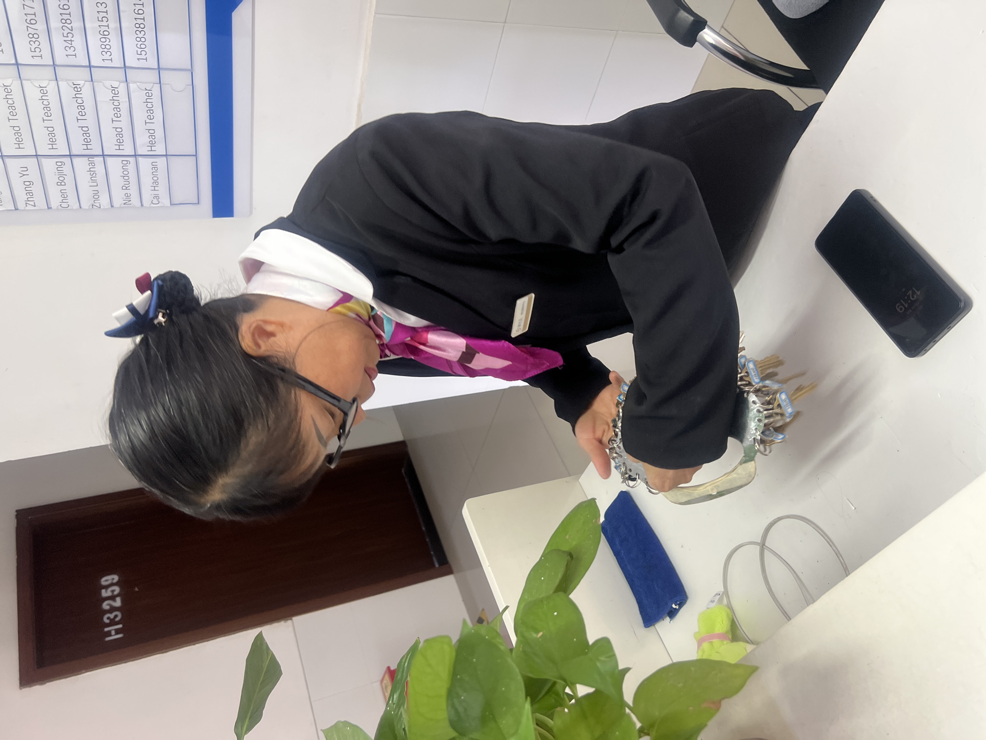 A woman in a black blazer with a white undershirt and a pink patterned scarf is pictured inside a building, likely an office or school setting, in what may be Jiangjin, China. Her hair is up in a bun with a blue and navy-blue hair accessory. She is wearing glasses and appears to be examining a key chain. A black smartphone is placed on the white desk to the right, displaying a time of 12:19. A potted plant with green leaves is in the foreground. The text "H3259" is visible on a brown door behind the plant. On the wall, there is a table listing names and titles under "Head Teacher" alongside numbers, possibly phone numbers. The names listed include Zhang Yu, Chen Bojing, Zhou Linshan, Nie Rudong, and Cai Haonan. The location is suggested as Jiangjin, China, based on the Chinese names and titles.