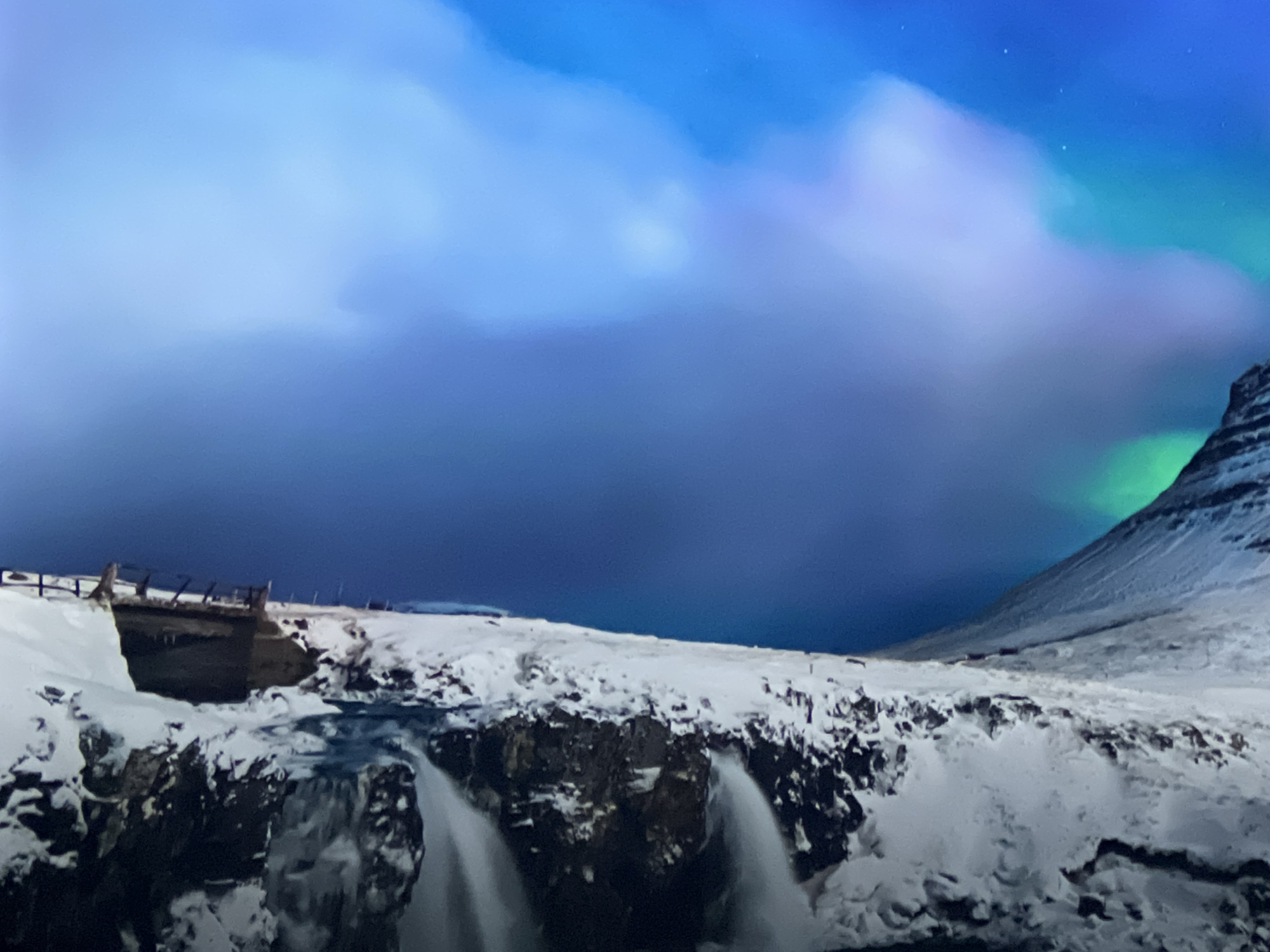 This media file depicts a dramatic winter landscape featuring a waterfall, a snow-covered mountain, and the aurora borealis. The waterfall cascades over a rocky, snow-dusted precipice, with its spray partially frozen. To the left of the waterfall, a wooden bridge or viewing platform with railings is visible, also covered in snow. The surrounding terrain is blanketed in thick snow, giving the scene a pristine, icy appearance.
Dominating the upper portion of the image is the sky, a canvas of swirling blues, purples, and greens. The vibrant colors and ethereal glow indicate the presence of the aurora borealis, also known as the Northern Lights. The aurora creates an otherworldly atmosphere, contrasting with the stark beauty of the frozen landscape below. A mountain, its slopes also covered in snow, rises on the right side of the frame, partially illuminated by the aurora.
The scene appears to be set at night or during twilight, given the darkness of the sky and the glow of the aurora. The weather is clearly cold and snowy. The overall mood is one of awe and wonder, capturing the raw, untamed beauty of nature in a polar region. There are no visible people or text in the image. The combination of the powerful waterfall, the majestic mountain, and the dancing lights in the sky creates a breathtaking and memorable spectacle.