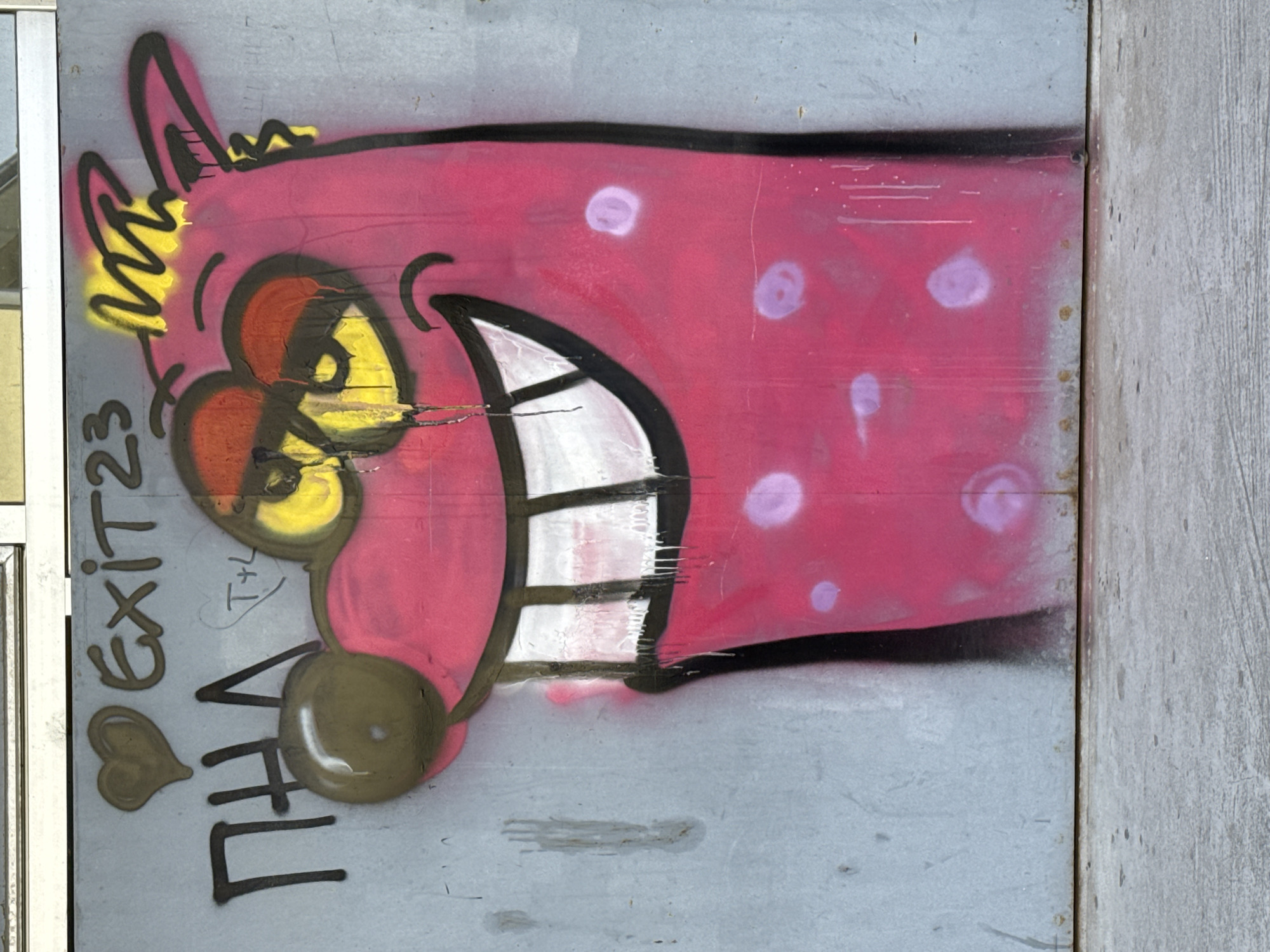 A piece of graffiti art is painted on a gray metal surface. The graffiti depicts a cartoonish pink creature with large teeth and purple spots. Beside the creature, graffiti reads "EXIT 23" and "I <3 THA." The location is Novi Sad, Serbia, but the specific location within the city cannot be determined from the image.