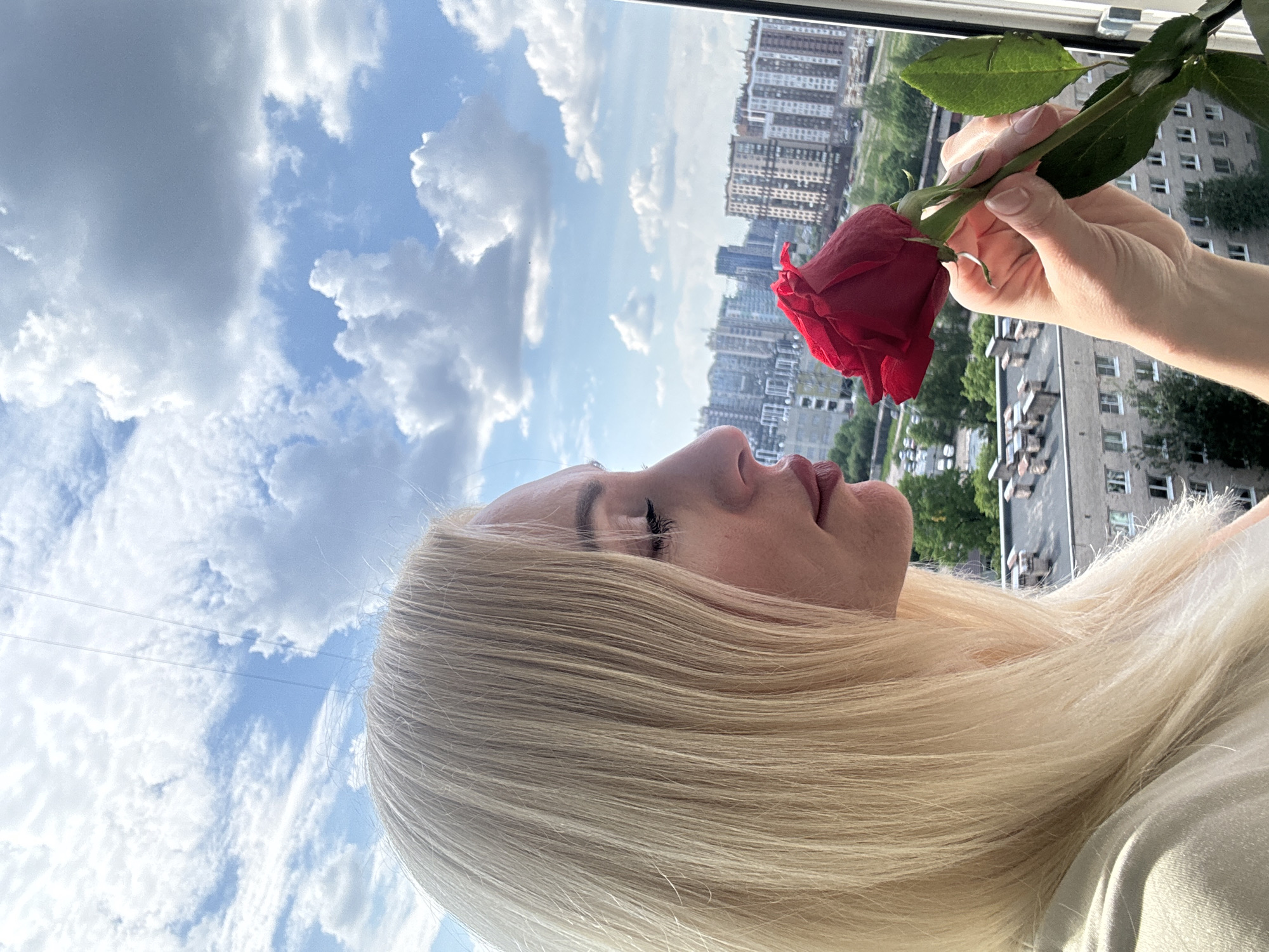An image captured from a tilted perspective shows a person holding a red rose to their face against a bright sky and urban backdrop. The horizon is oriented vertically on the right side of the frame, with the sky filling the left.
The primary subject is a person, likely female, with long, flowing blonde hair, viewed in profile. Their head is tilted back, with eyes seemingly closed or looking down, and their lips slightly parted. They are bringing a vibrant red rose, held by their right hand with neatly manicured nails, close to their nose, appearing to inhale its scent. The person is wearing a light-colored top, partially visible at the bottom right.
In the background, a vast, clear blue sky dominates, interspersed with numerous fluffy white clouds, indicating a bright and sunny day. Beyond a window frame, an urban landscape unfolds, featuring various high-rise and mid-rise residential buildings. Some green foliage from trees is also visible among the structures, suggesting a city environment.
The scene conveys a moment of calm and sensory focus, as the individual appears immersed in the fragrance of the rose, contrasted with the bustling city outside. There is no visible text in the image.