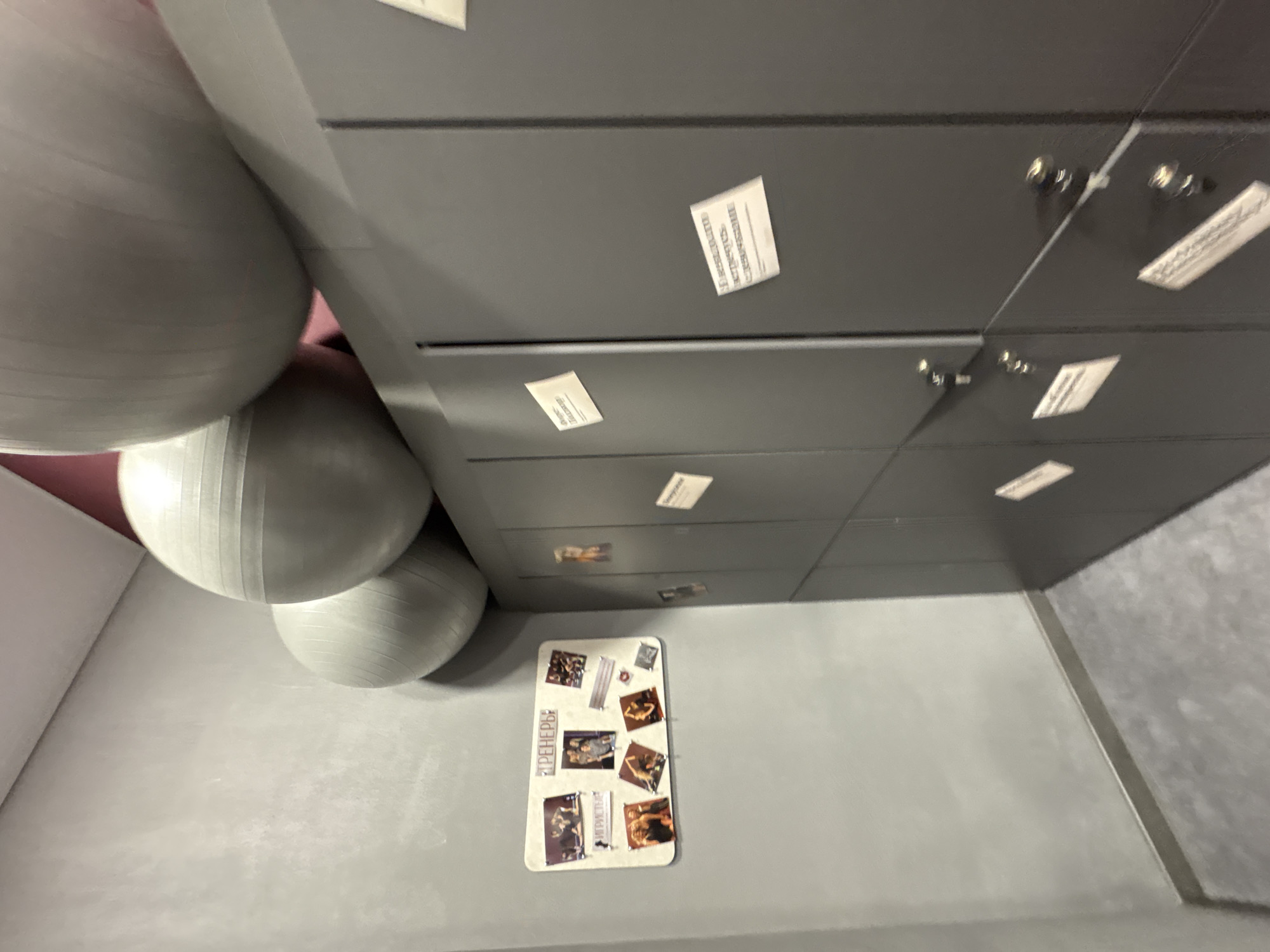The image shows a room with exercise balls and lockers. Three gray exercise balls are stacked on top of each other on the left side of the frame. Adjacent to the balls is a bank of gray lockers with small white labels or notices affixed to each locker. A rectangular sign with pictures and Russian text is attached to the wall near the base of the lockers. The walls are painted a light gray color. Based on the presence of Russian text and online searches, the image likely originates from a gym in Izhevsk, Russia, possibly the Fitness House gym.