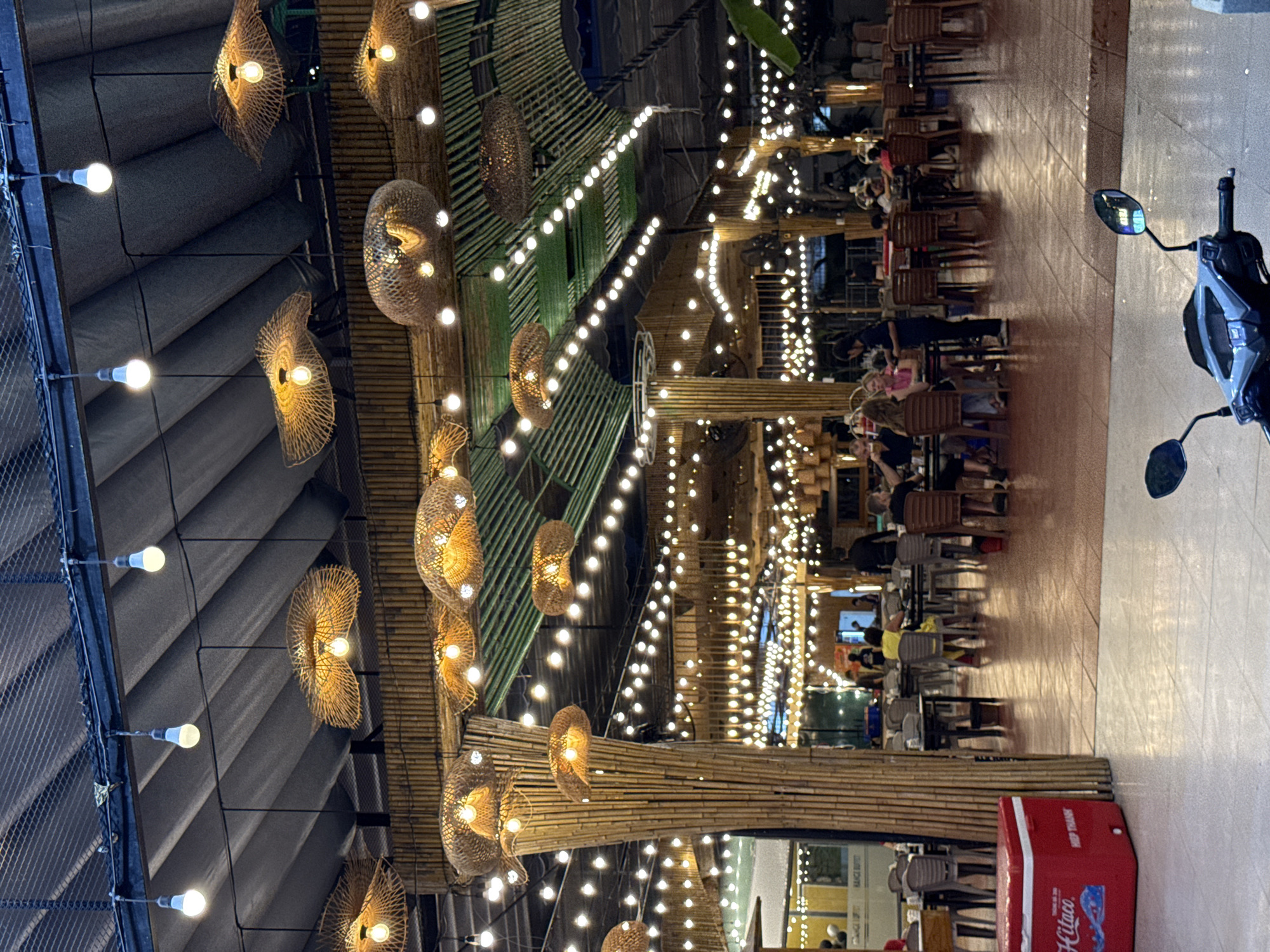 The image depicts an outdoor dining area, likely a restaurant, characterized by a thatched roof supported by bamboo columns. Numerous woven light fixtures hang from the ceiling, casting a warm glow. String lights are also visible, adorning the overhead structure. Several tables and chairs are arranged throughout the space, with some occupied by patrons. In the foreground, a red cooler is placed on the tiled ground, and a motorcycle is partially visible. The scene suggests a relaxed evening atmosphere, possibly in a coastal or tropical setting. While the specific location in Nha Trang, Vietnam, cannot be pinpointed from the image alone, the architectural style and ambiance are consistent with many establishments in such a region.