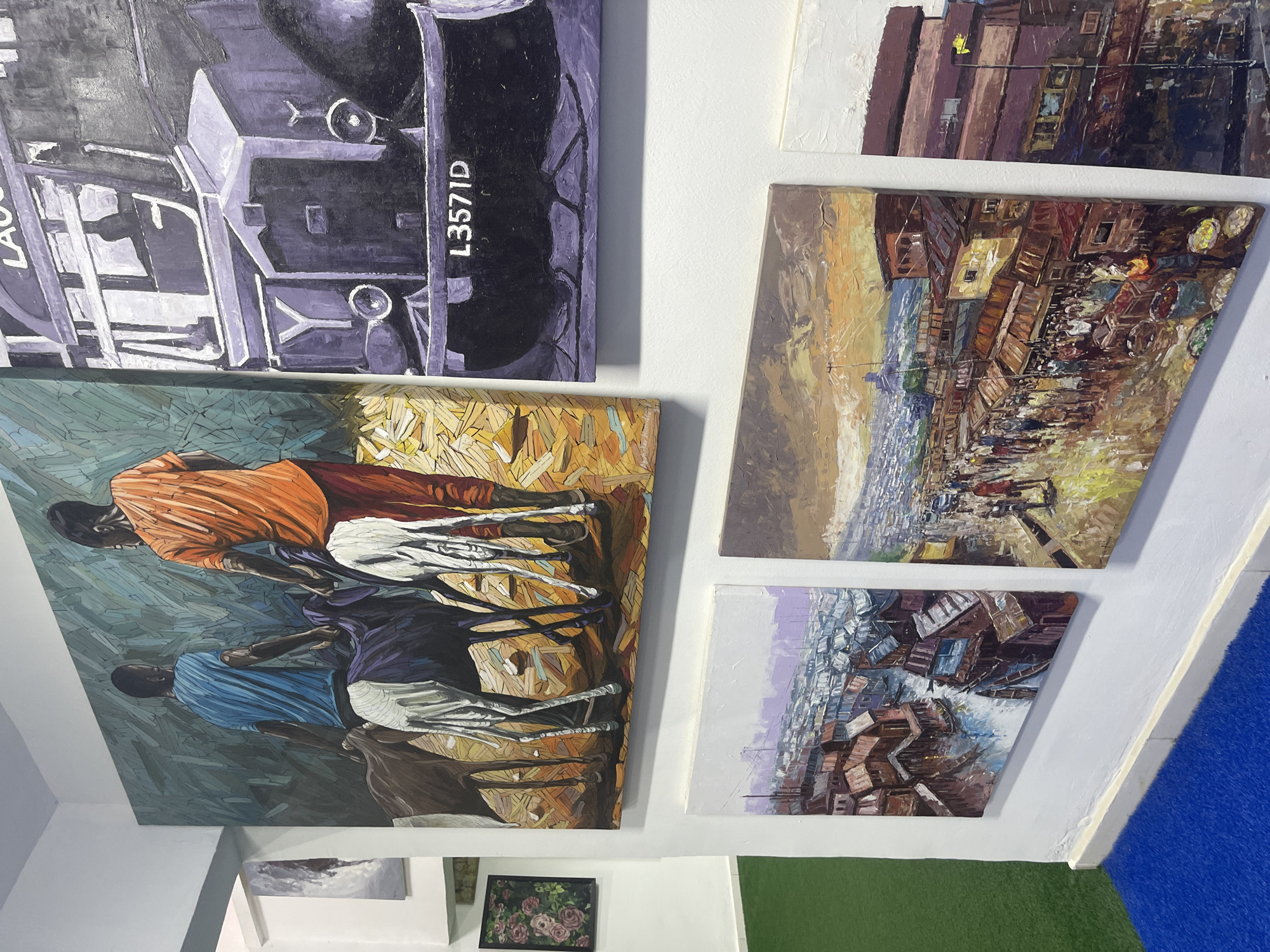 The image displays an assortment of paintings on white walls, characteristic of an art gallery or exhibition space in Abuja, Nigeria. The photo is taken at an angle, resulting in several artworks appearing rotated.
The largest painting, on the left, depicts two dark-skinned male figures. One, in an orange top, stands next to a white goat, while the other, in a blue top, is beside a dark brown goat. The ground and background are rendered with distinct, geometric, and textured patterns in shades of green, yellow, and brown. The figures and animals are painted with a stylized, almost fragmented aesthetic.
Above this, a horizontal painting features a large, dark purple and black vehicle, possibly a bus or truck. Visible text on its side includes "L3571D" and a partial "LAUS," likely indicating "LAGOS."
On the right side, multiple smaller paintings primarily portray vibrant urban landscapes or market scenes. These artworks show densely packed buildings, street activity, and crowds, utilizing expressive, textured brushstrokes. One painting employs warm tones of brown, orange, and yellow, suggesting a daytime or sunset setting, while another uses cooler purples and blues for a similar cityscape.
At the bottom of the wall, a small, framed abstract painting with muted tones and a small framed picture of pink and red roses are also visible. The floor features a blue carpet and green artificial grass, completing the exhibition setting. The overall lighting is bright, highlighting the diverse subjects and artistic styles.