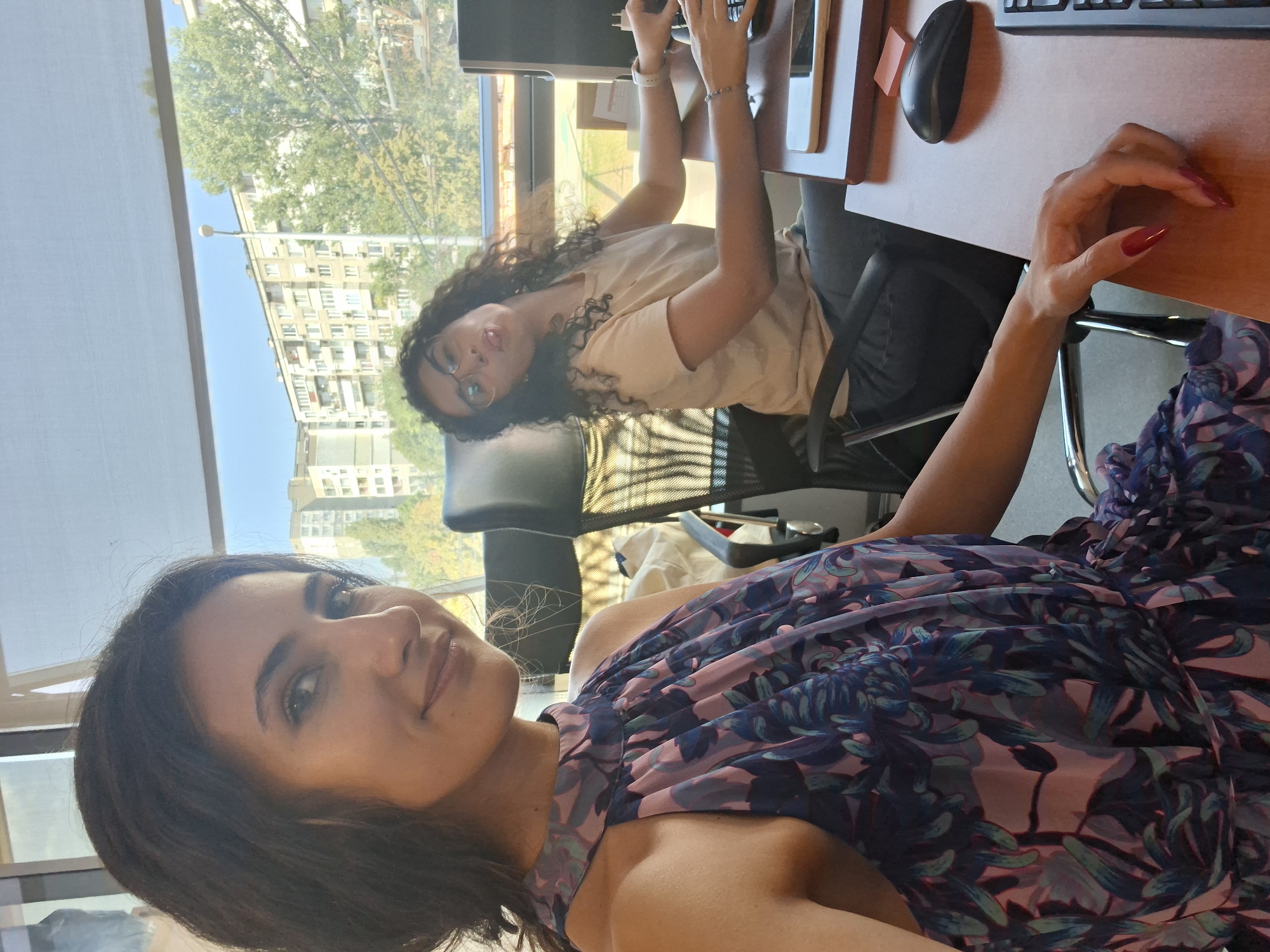 Two young women are in an office environment, likely a shared workspace, with a large window overlooking a city. The woman in the foreground, on the left, is looking directly at the camera and smiling. She has long, dark, wavy hair and is wearing a floral-patterned halter-neck dress in shades of blue and purple. The woman in the background, on the right, is at a desk and is making a playful face, sticking her tongue out. She has curly dark hair and is wearing glasses and a light-colored t-shirt. She is seated at a desk with a computer keyboard and mouse, and a mobile phone. The office appears to be well-lit by natural light streaming in from the window, which offers a view of apartment buildings and trees.