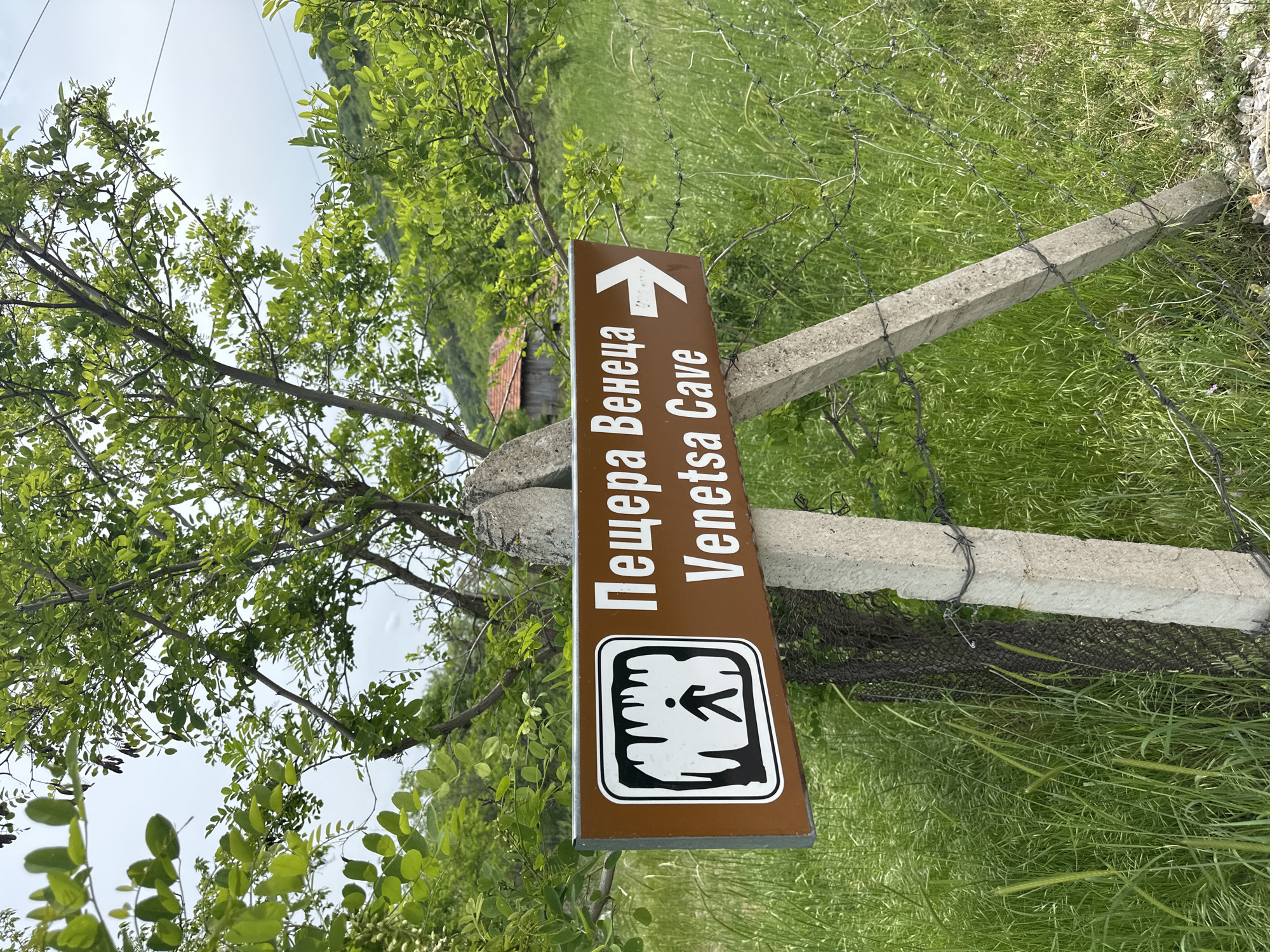 A sign for "Venetsa Cave" in Gara Oreshets, Bulgaria, is attached to two concrete posts with barbed wire. The sign is brown with white lettering and a white arrow pointing upwards. Below the text, a symbol depicts a walking figure. The area surrounding the sign is grassy with some trees and a glimpse of a building in the background. The sky is overcast.