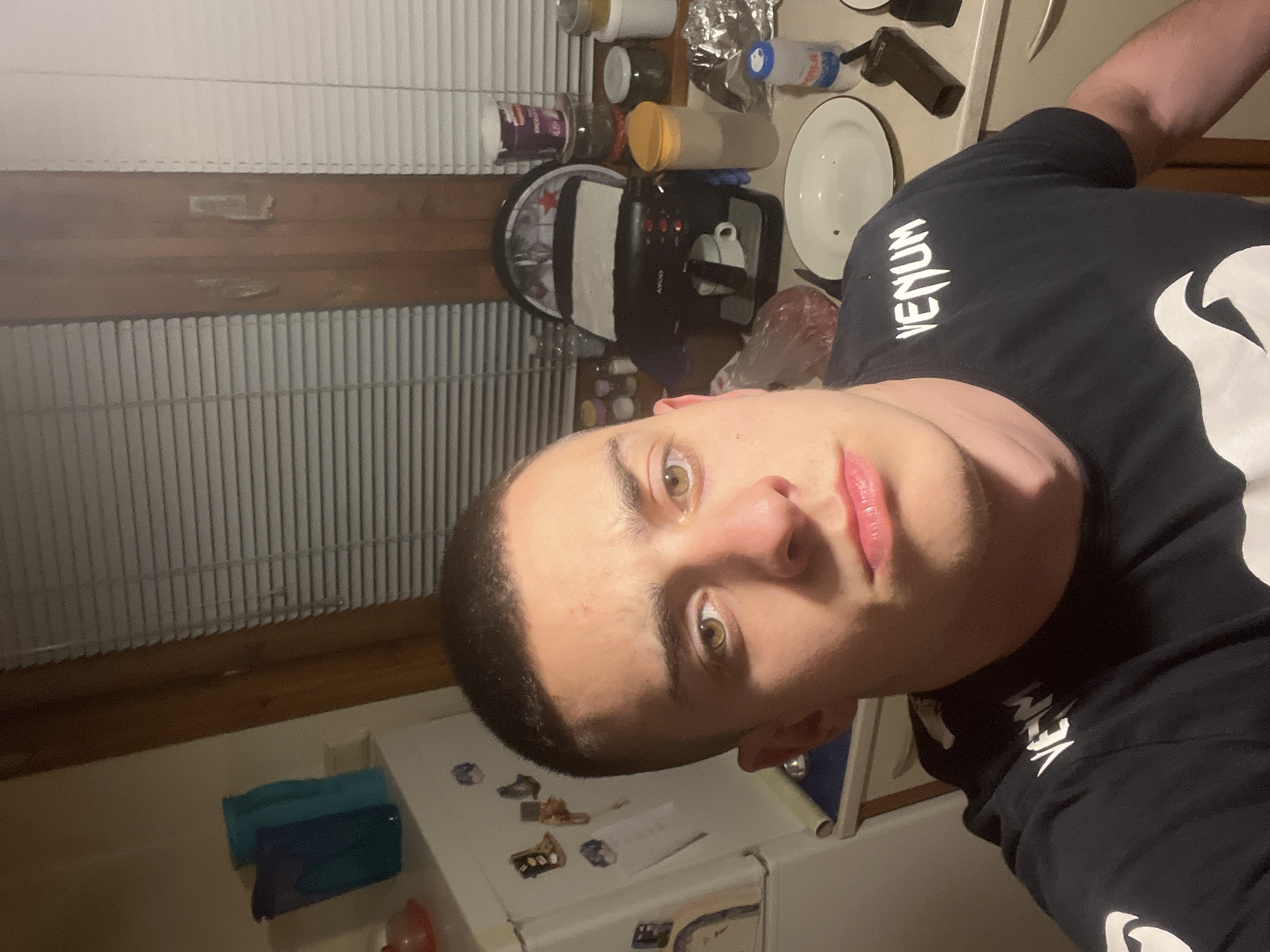 A selfie taken indoors, in what appears to be a kitchen or pantry. The subject is a fair-skinned male with short, dark hair. He is wearing a black t-shirt with a white logo and the word "VENUM" visible on the upper left side of the shirt. His expression is neutral, and he is looking directly at the camera.
The background reveals a cluttered environment. There is a window with blinds to the left, and a countertop with various items, including containers, a plate, and what appears to be a coffee maker. A white refrigerator with magnets is below the subject. The location is impossible to ascertain based on the available information.