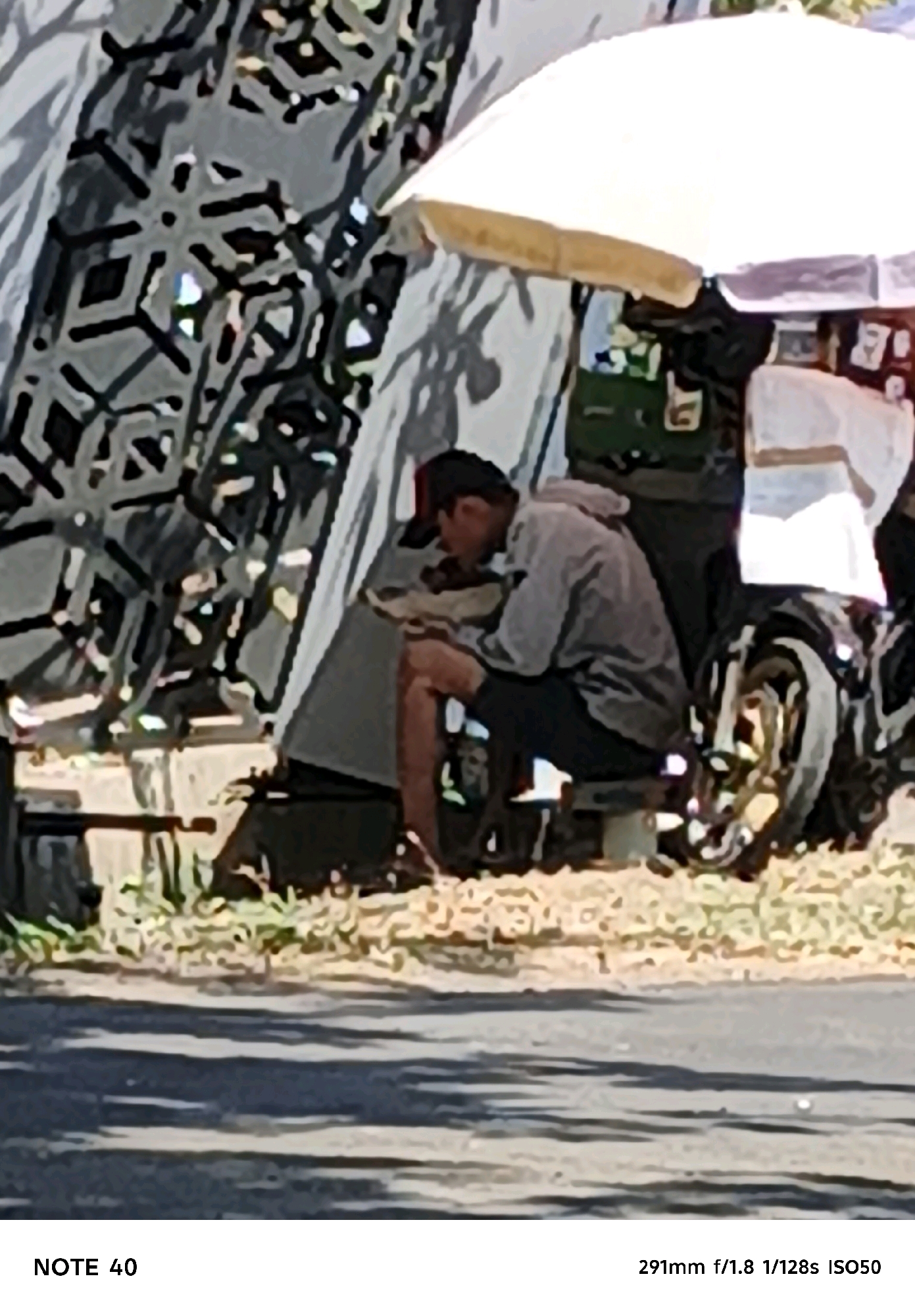 A person sits on a bench under a white umbrella at a roadside stall. They are wearing a grey hoodie and dark pants, and appear to be eating or looking at something in their hands. A motorcycle is parked behind them. The stall has various items displayed, including what looks like drinks in bottles and packets. The background features a patterned wall or fence and foliage. The ground is paved with asphalt, and there is dry grass and dirt around the stall. The lighting suggests it is daytime. The image includes metadata indicating the camera settings: 291mm f/1.8 1/128s ISO50. The text "NOTE 40" is present at the bottom left.