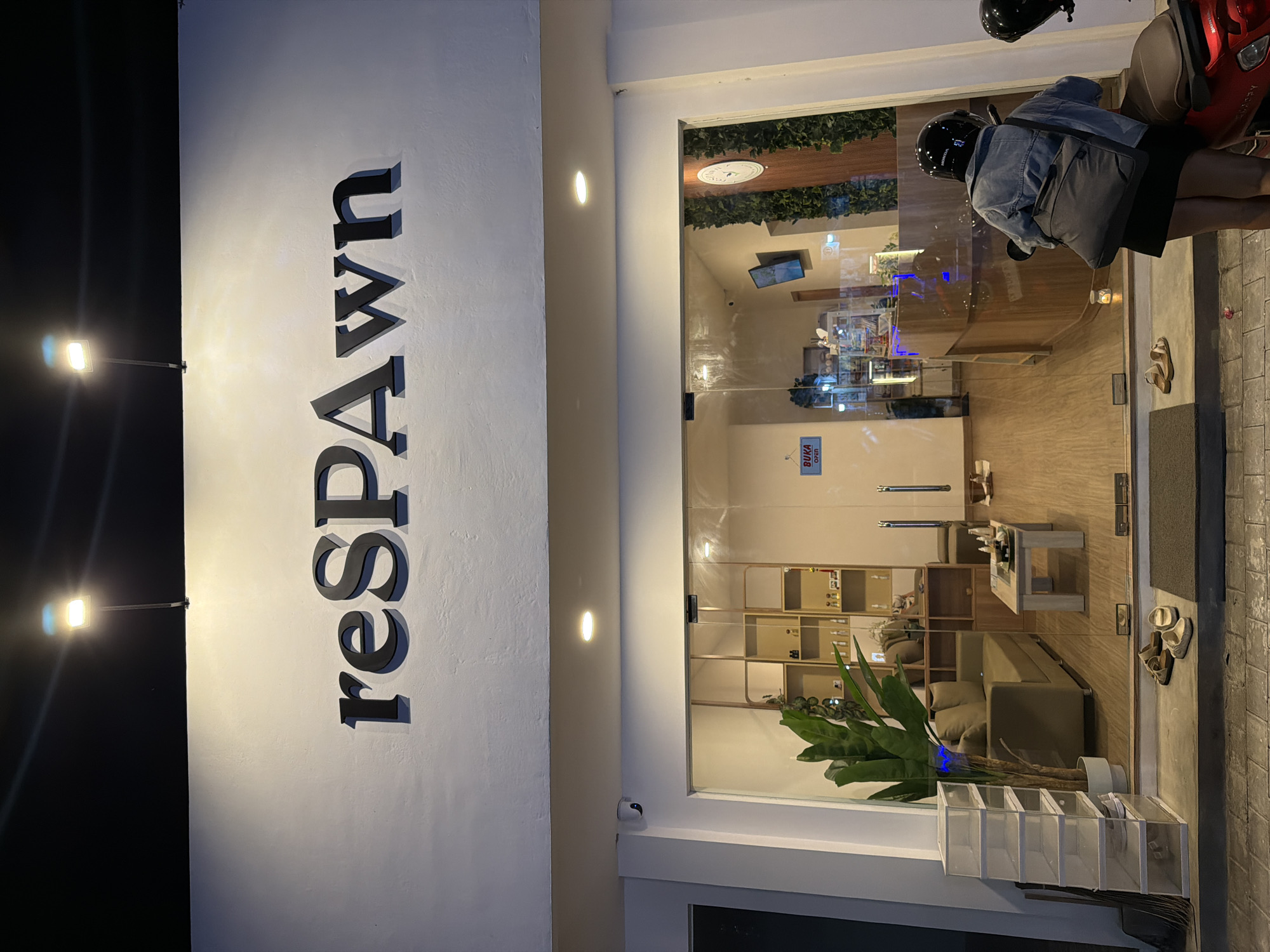 The image shows the exterior of a business named "reSPAWN" located in North Kuta, Indonesia. The name is vertically oriented on a white wall to the left. To the right is a glass entrance showcasing the interior, which includes shelving, furniture, and a potted plant near the entrance. A person wearing a helmet and carrying a bag is near a red scooter parked just outside the entrance. Streetlights illuminate the scene. The sidewalk outside the entrance is visible, with a few pairs of sandals.