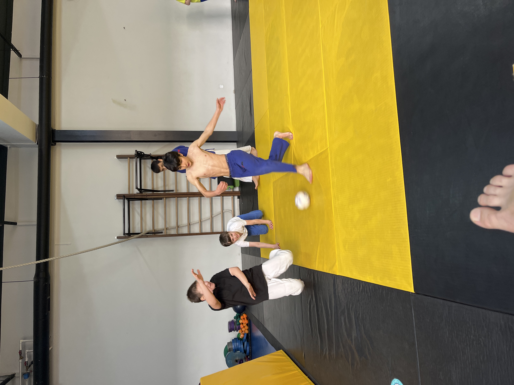The image shows a group of people in what appears to be a training facility. It's challenging to confirm the location as Krasnoyarsk, Russia, without additional information or context.
The scene includes:
* Several individuals engaged in physical activities on a mat.
* One shirtless person in blue pants is in mid-air, possibly kicking a ball.
* Another person is kneeling on the mat.
* A person in black attire is on the mat with their hands raised.
* The training facility environment features a wall ladder, mats of different colors, and a rope hanging from the ceiling.
* There are also some weights and equipment visible in the background.