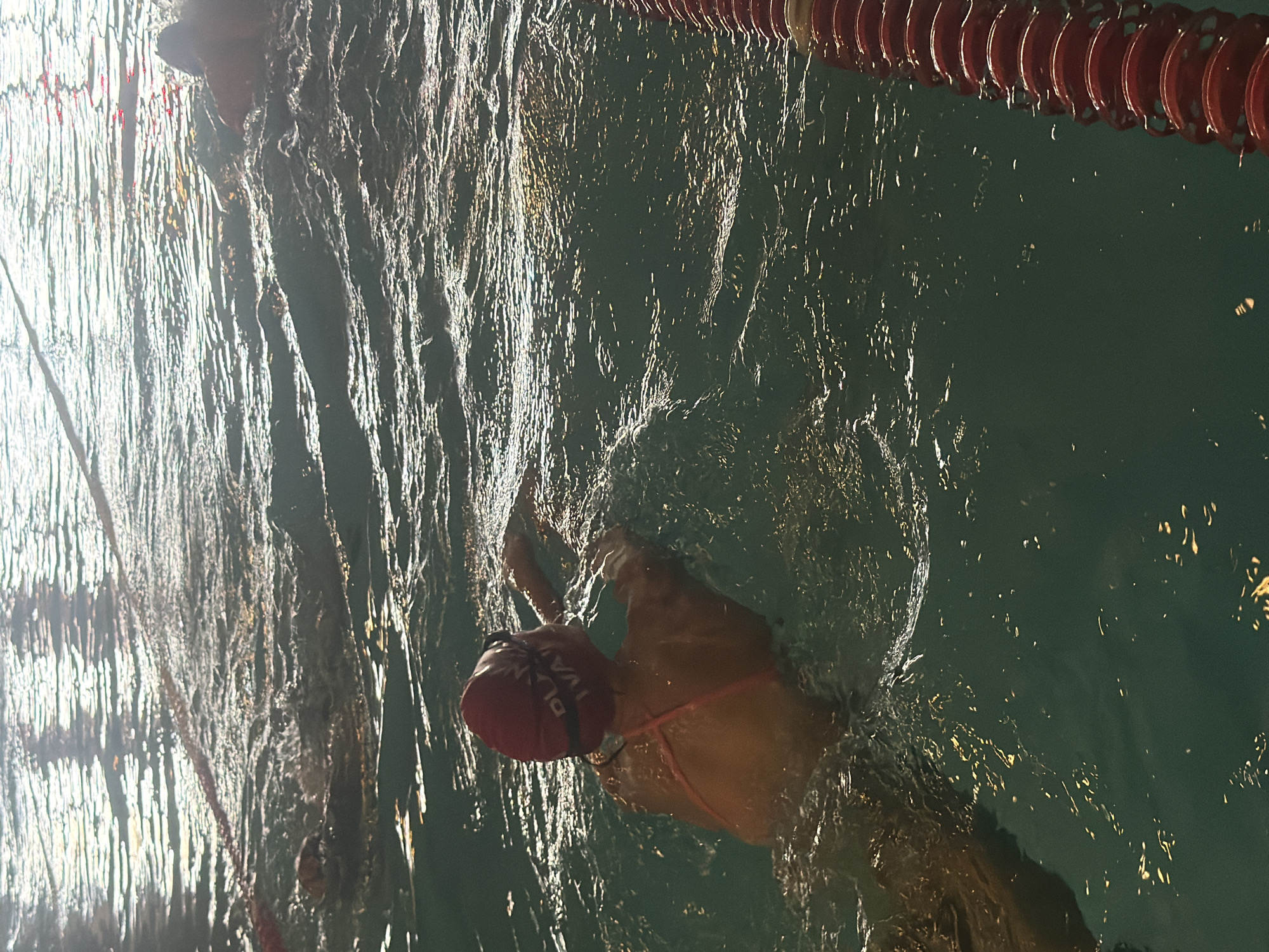 The image shows a swimmer in a swimming pool. The swimmer is wearing a red swimming cap with white lettering and black goggles. The swimmer is partially submerged in the water, with their upper body and head visible. The water is rippled and reflects the overhead lights. Lane lines are visible in the water, and a red lane rope is on the right side of the image. The scene appears to be an indoor swimming pool, possibly at night or in low light conditions. There is no information in the image to suggest a specific location in Kyiv, Ukraine.