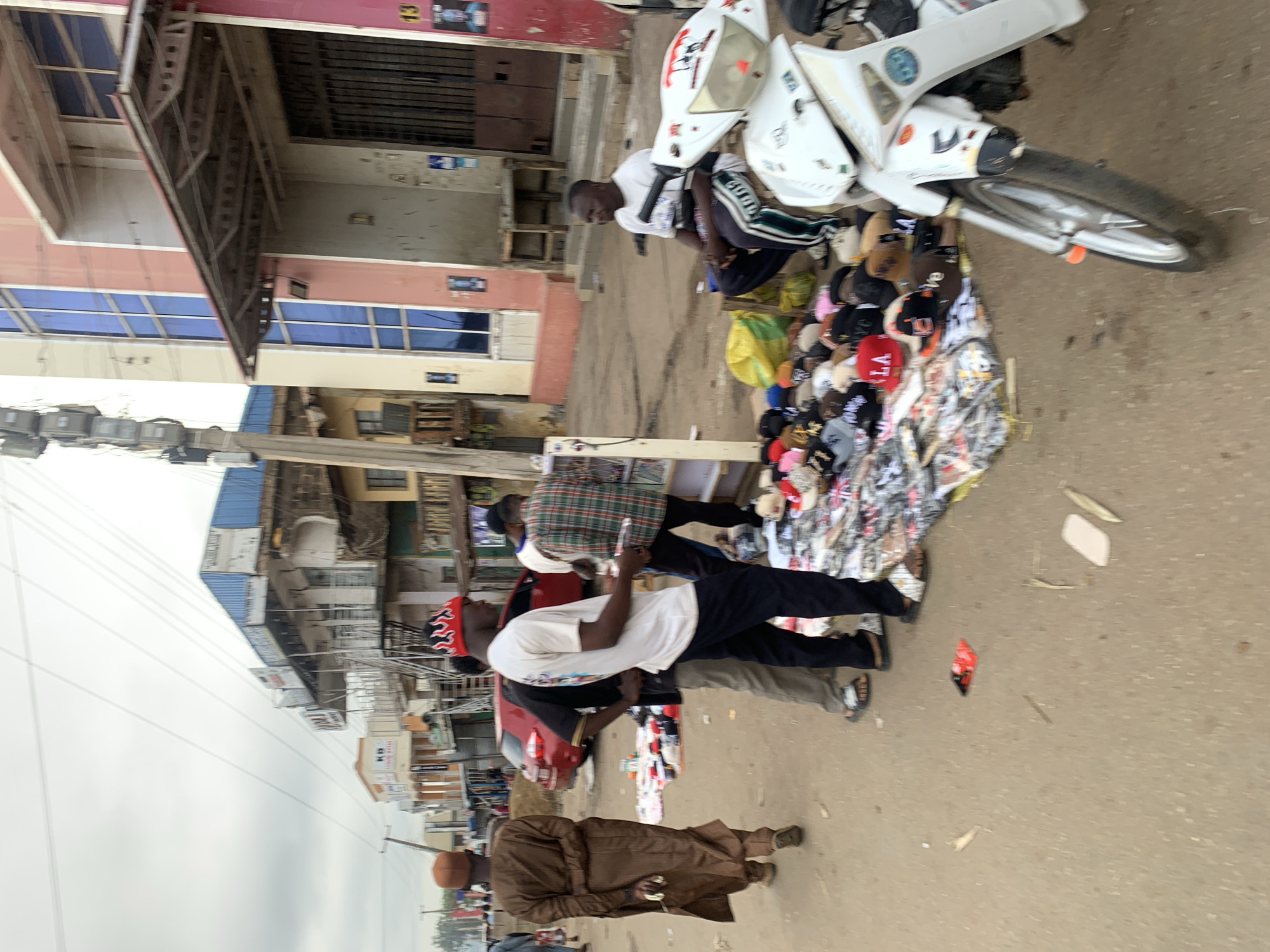 A street scene in Jos, Nigeria features several people and a variety of objects. In the foreground, a man in a brown robe walks along the side of the road. Several people stand near a display of hats and other items laid out on a cloth on the ground. One person is perched on a white motorcycle. The backdrop includes buildings of varying heights and architectural styles, with a power line visible in the sky. The weather appears overcast.