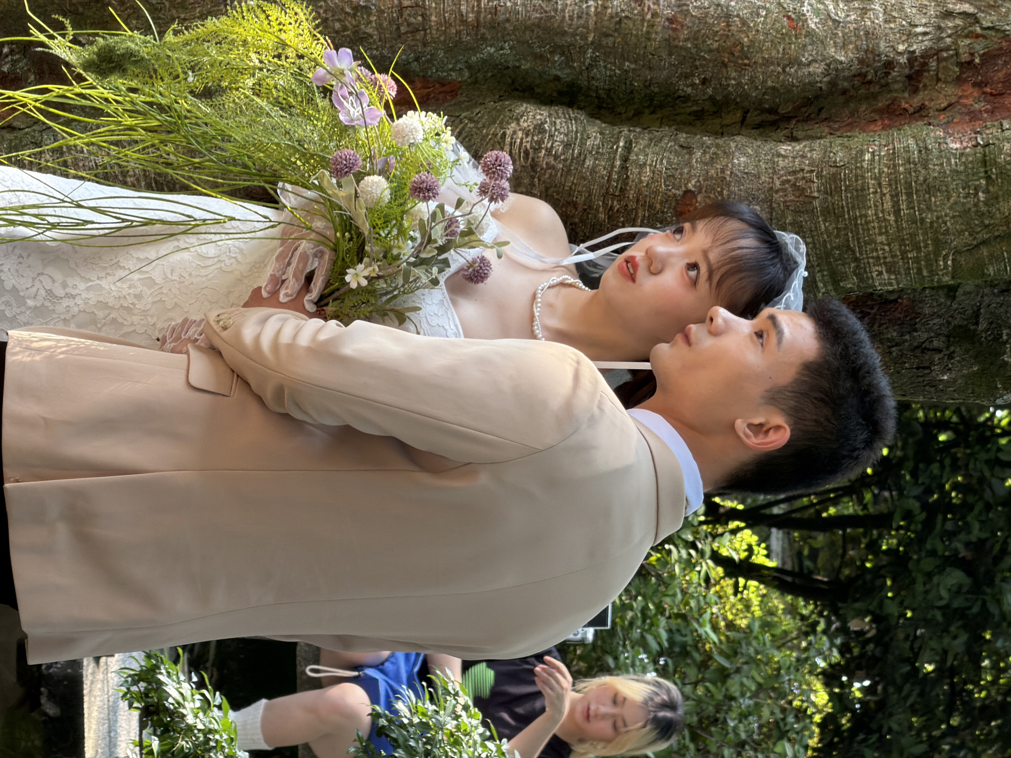 In a verdant outdoor setting, likely within Jiangbeicheng Street, China, a bridal couple is the central focus of the photograph. The bride, positioned to the left, wears a white lace wedding dress and a delicate veil, clutching a bouquet of assorted flowers. She is adorned with a pearl necklace. Beside her, the groom is attired in a light-colored suit jacket over a white collared shirt. Their gaze is directed off-camera to the right, suggesting an interaction or observation outside the frame. In the background, a young woman with blonde hair sits on a stone bench, dressed in a black t-shirt and blue shorts, with white socks. The scene is framed by the textured bark of a large tree on the left and lush green foliage that drapes across the foreground and background, indicating a natural, perhaps park-like environment. Sunlight filters through the leaves, creating dappled light across the subjects and the surroundings.