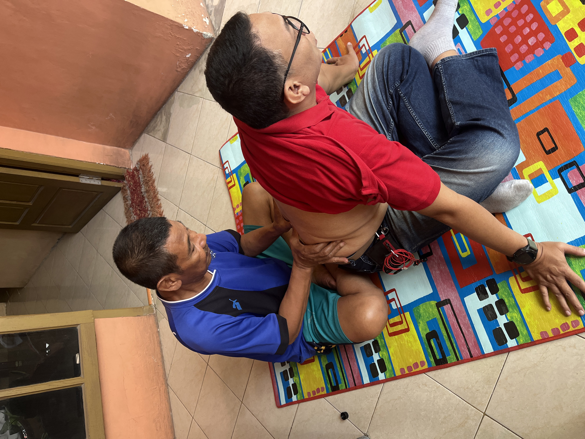 Two adult males are on a colorful mat placed on a tiled floor, likely in front of a residential building in the Sidorukun area of Indonesia. The man on the left, wearing a blue shirt with a black collar and turquoise shorts, is seated and appears to be giving a massage to the man on the right. The man on the right is wearing a red polo shirt, blue denim jeans, and grey socks. He is lying on his stomach, with his head turned to the side. His back is exposed, and the man on the left is using his hands to massage his midsection. A black smartwatch is visible on the right man's left wrist, and red wires connected to what appears to be a device are clipped to the right man's waistband. The environment suggests a casual outdoor or semi-outdoor setting, with a portion of an orange-colored wall and a doorway visible in the background. The tiled floor extends beyond the mat, and a small rug is placed near the doorway.