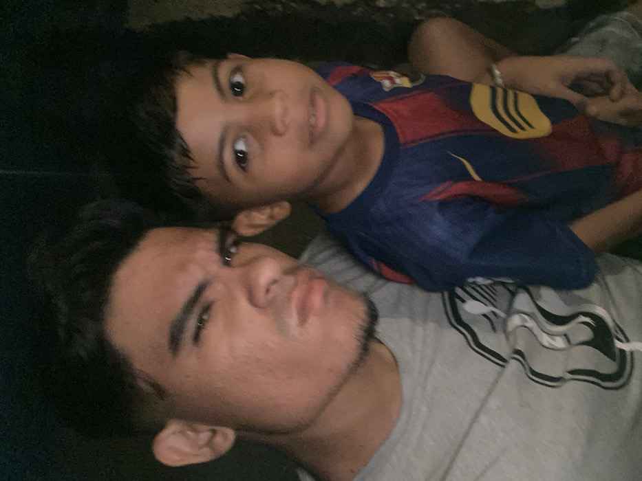 A man and a young boy are lying down, likely together. The man has short, dark hair and a slight beard and mustache. He is wearing a grey t-shirt. The boy is wearing a blue and red soccer jersey with yellow accents. They are close together and both looking directly at the camera. The lighting is dim, creating some shadows.