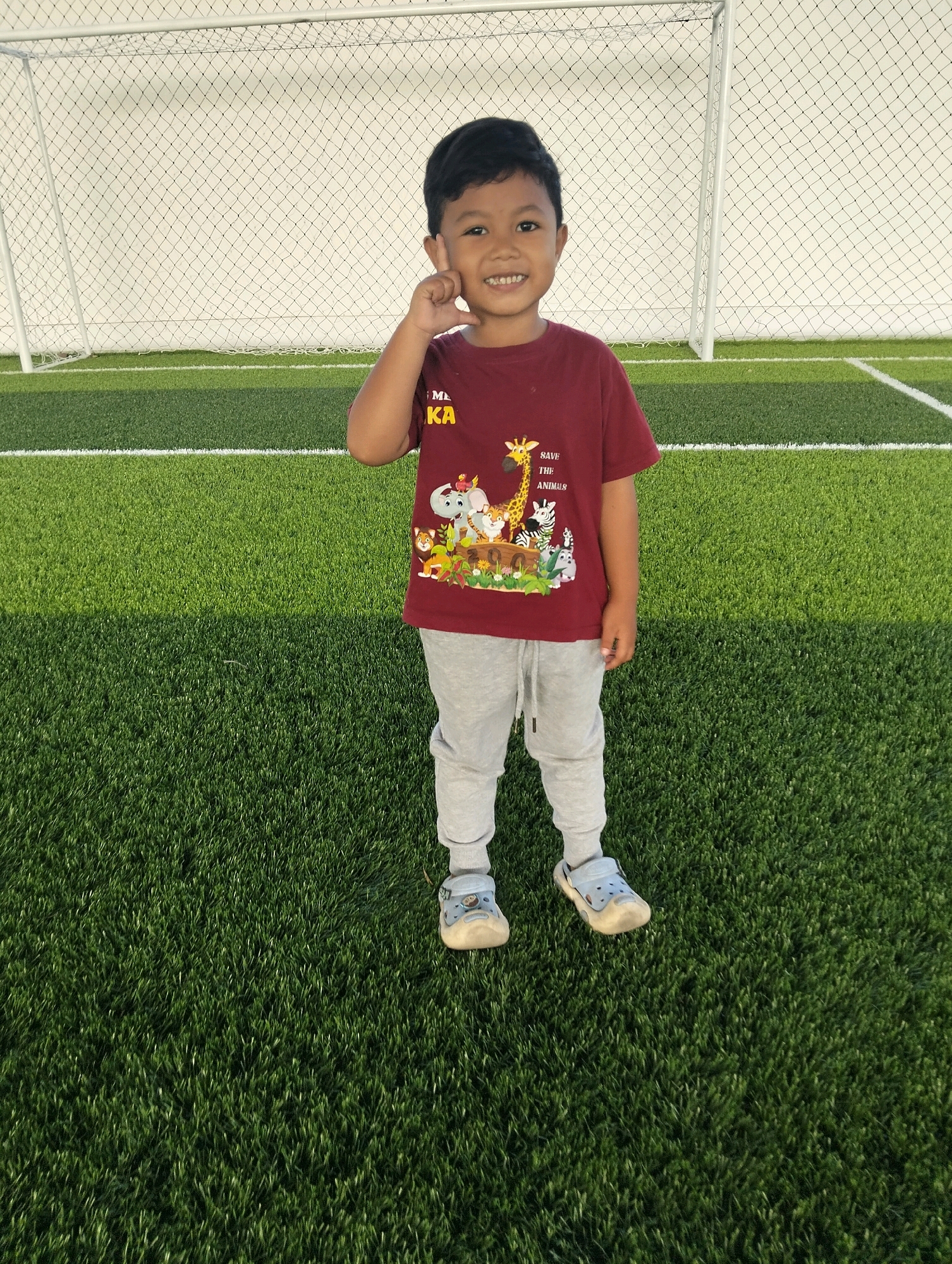 A young boy with short black hair is standing on a bright green artificial turf field. He is smiling and has his right index finger touching his right cheek. He is wearing a maroon t-shirt with an animal graphic and the words "SAVE THE ANIMALS" printed on it, along with light gray sweatpants and blue slip-on shoes. In the background, a white soccer goal with a black net is visible. The field appears to be part of a sports complex. The location is identified as Solokuro, Indonesia.