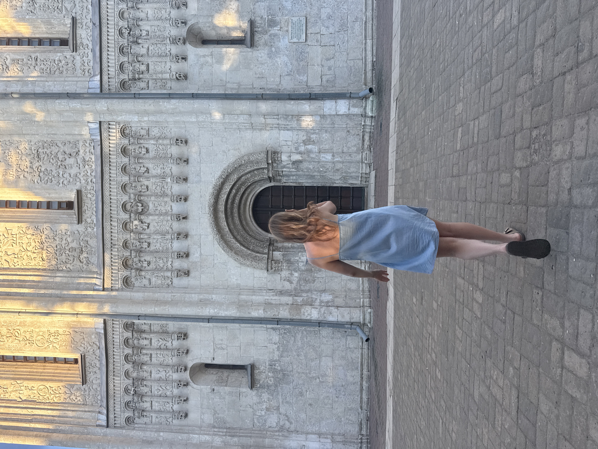 A woman with blonde hair wearing a light blue dress and dark sandals walks away from the camera towards an ornate stone entrance of a large, ancient building. The building's facade is intricately carved with decorative patterns and figures, and it is bathed in warm, late afternoon sunlight. The ground in front of the building is paved with large, rectangular stones. The scene appears to be in front of the Dormition Cathedral in Vladimir, Russia, identifiable by its distinctive white stone architecture and elaborate carvings.