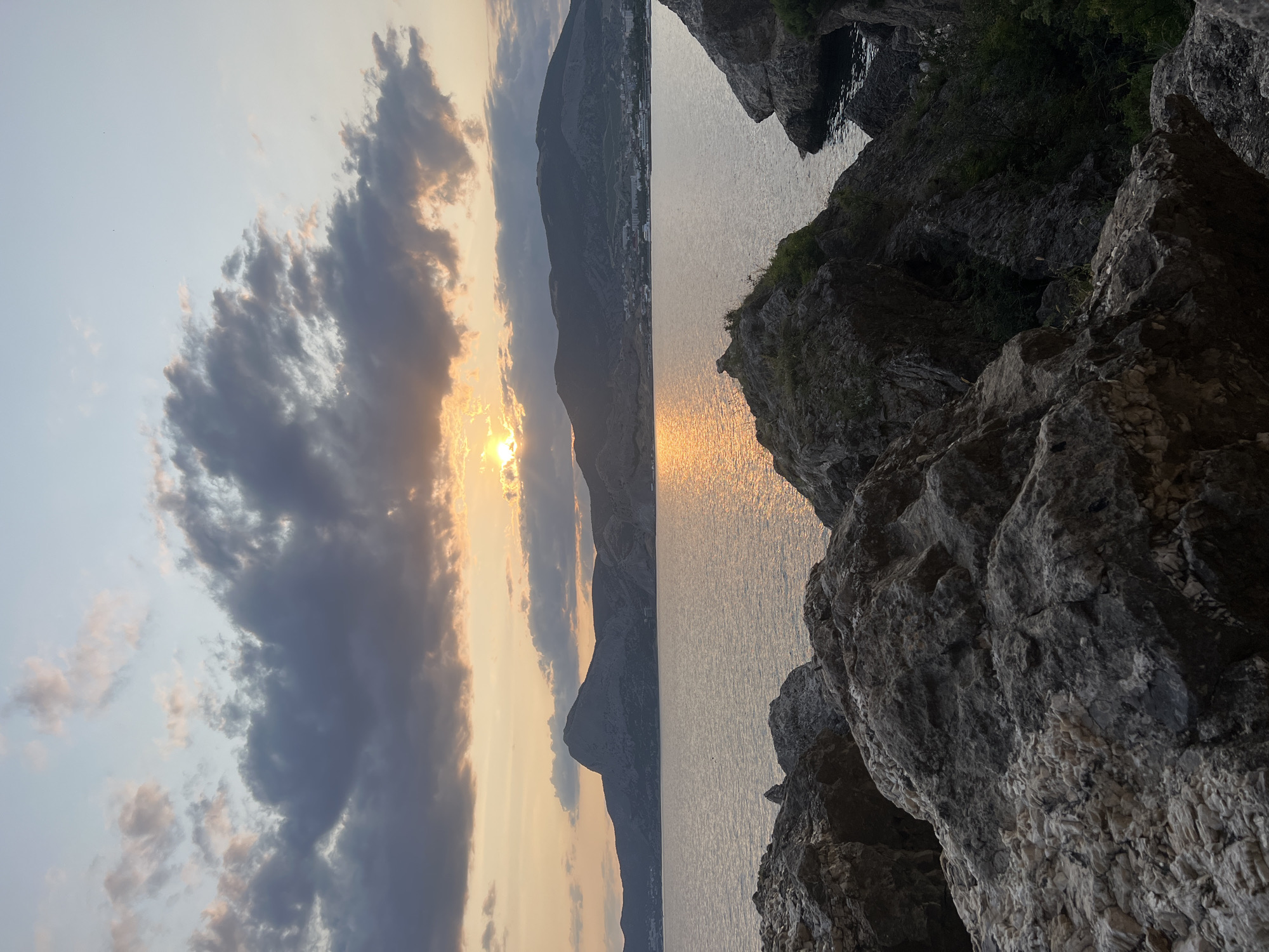 The image depicts a coastal landscape at sunset, likely near Sudak, Ukraine, given the rocky shoreline and mountainous backdrop. In the foreground, large, weathered rocks are prominent, textured with lighter and darker shades, suggesting erosion and age. Some sparse green vegetation can be seen growing amongst the rocks.
Beyond the rocks lies a calm body of water, reflecting the warm hues of the setting sun. The water surface is gently rippled, creating shimmering pathways of light.
In the middle ground, a range of mountains stretches across the horizon. The mountains are a muted gray and brown, with distinct peaks and slopes. A settlement, with buildings clustered together, is visible on the lower slopes of the mountains.
The sky above is a dramatic mix of colors. The sun, partially obscured by dark, wispy clouds, casts a golden and orange glow across the scene. The clouds themselves are a mix of dark grays and lighter, sunset-tinted whites and pinks, indicating the transitional light of dusk. The overall atmosphere is serene and tranquil. There are no people or animals visible in the image.