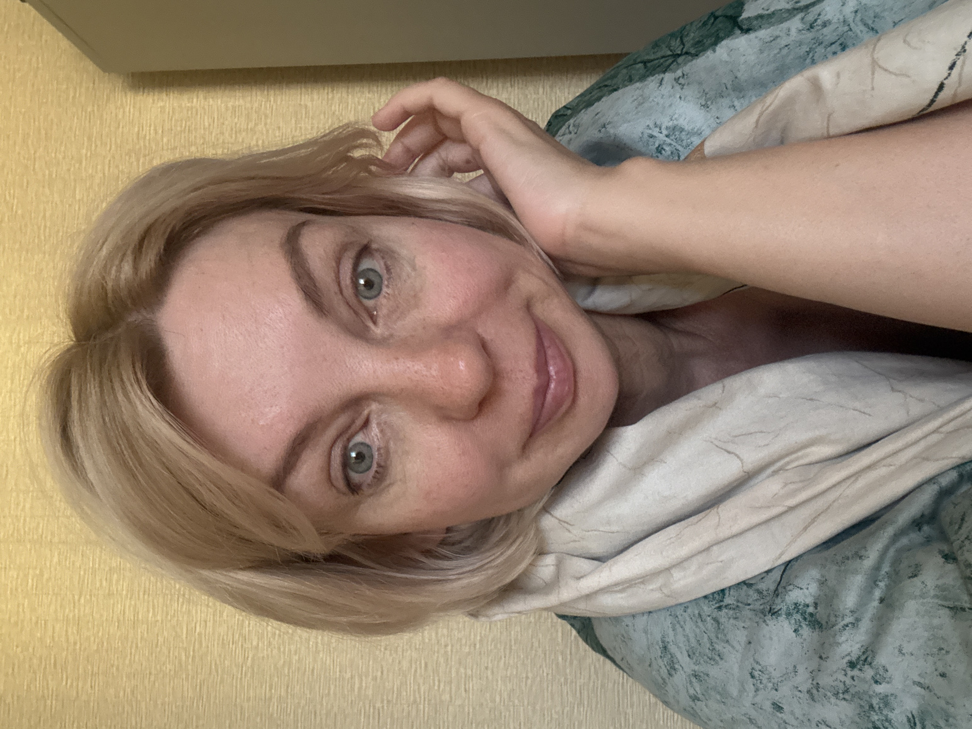 The image is a close-up, selfie-style photograph of a light-skinned woman with blonde, shoulder-length hair. She has blue eyes and is wearing a light-colored scarf and a patterned green garment. Her right hand is touching her face, resting near her ear. The background appears to be a wall with a textured, yellow wallpaper. There is a portion of a light-colored cabinet visible on the far right of the image. The lighting suggests an indoor setting. The image does not contain any identifiable landmarks or features that would indicate the city of Sochi, Russia, or any specific urban location.