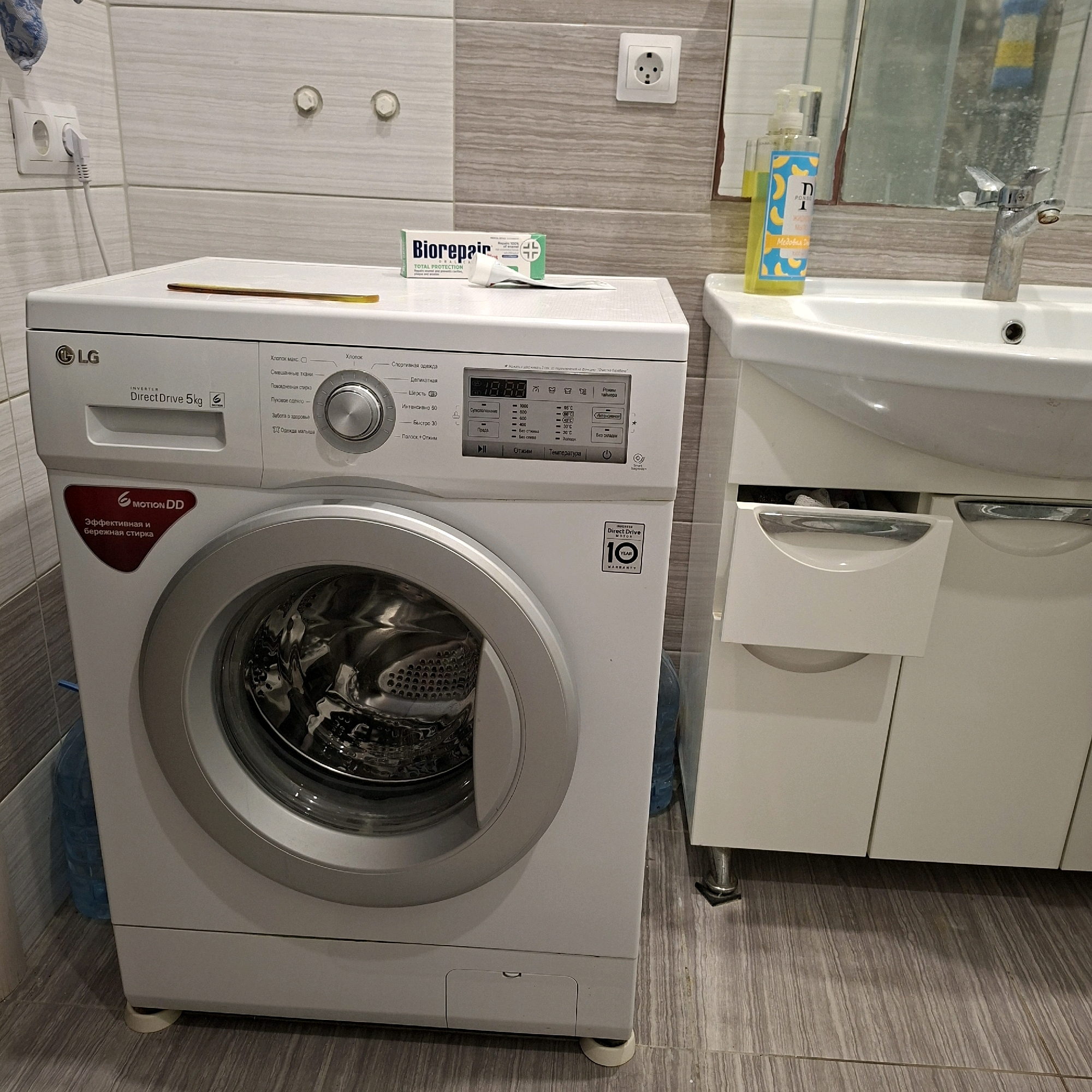 An LG washing machine, model DirectDrive 5kg, is positioned in a bathroom with tiled walls and a tiled floor. A sink with a faucet is located to the right of the washing machine. On top of the washing machine, a tube of Biorepair toothpaste is placed. A bottle of liquid soap with a banana motif is on the sink's edge. A power outlet is visible on the wall. The scene appears to be in a residential bathroom in Anapskaya, Russia.