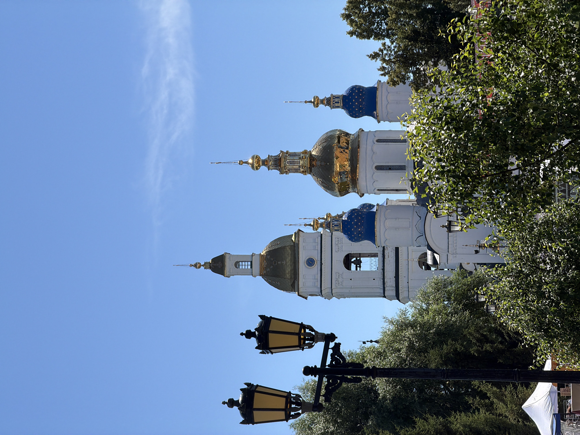 An outdoor, low-angle, medium shot shows a close-up of a black lamppost with two yellow lanterns on the left side of the frame, partially obscuring a portion of a white building with gold and blue domes in the background. The building appears to be a religious structure with multiple towers topped with golden spires and blue domes. A tall white bell tower with a clock face is visible to the left of the domes. Lush green trees and foliage are in the foreground and between the lamppost and the building. The sky above is a clear, bright blue with a few wispy clouds. A white tent is visible in the lower left corner of the image. The scene is brightly lit, suggesting a sunny day.