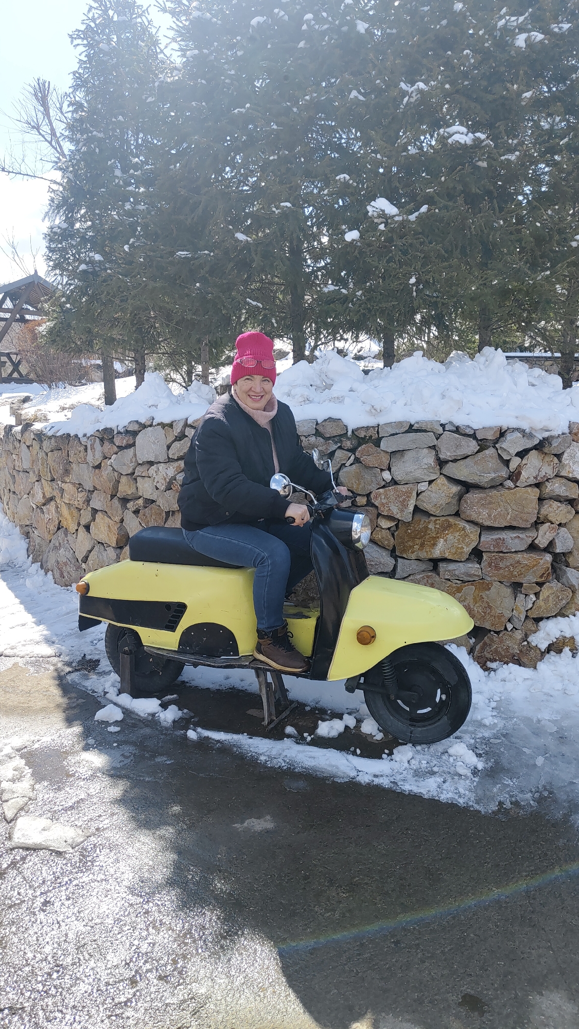 In an outdoor setting in Unknown, Unknown, an adult woman wearing a pink beanie, a black jacket, blue jeans, and brown shoes sits atop a yellow scooter. The scooter is parked on a damp paved surface, and the surrounding area shows remnants of snow. Behind the woman and the scooter is a stone wall with snow piled on top, and further back are evergreen trees. The woman is smiling and looking directly at the viewer.